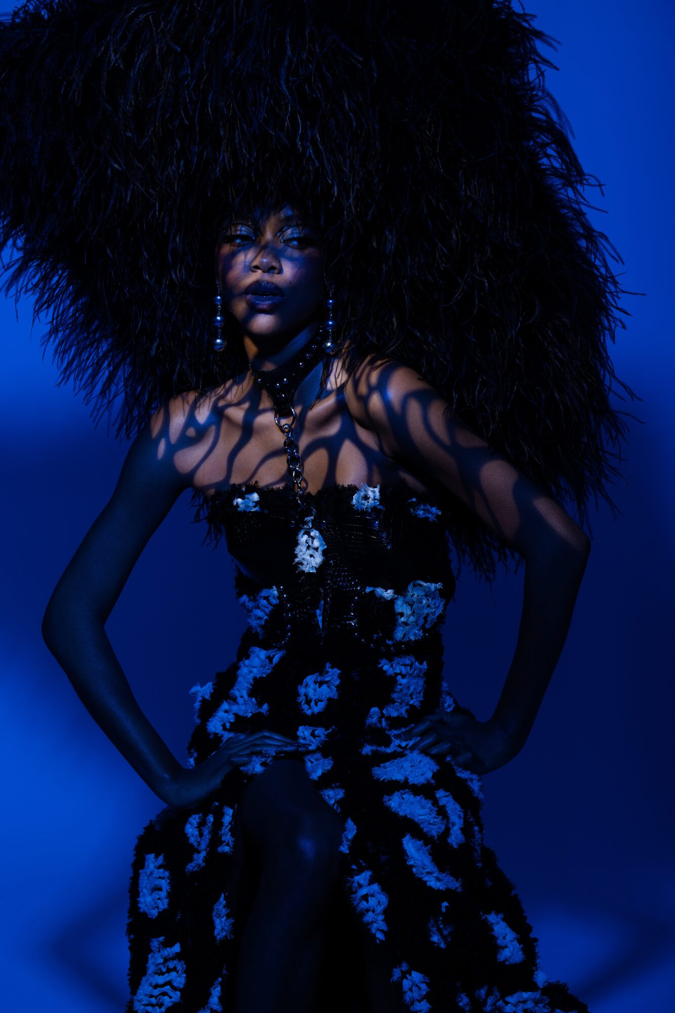Woman with large, textured hair and patterned dress, standing with hands on hips against a dark blue background.