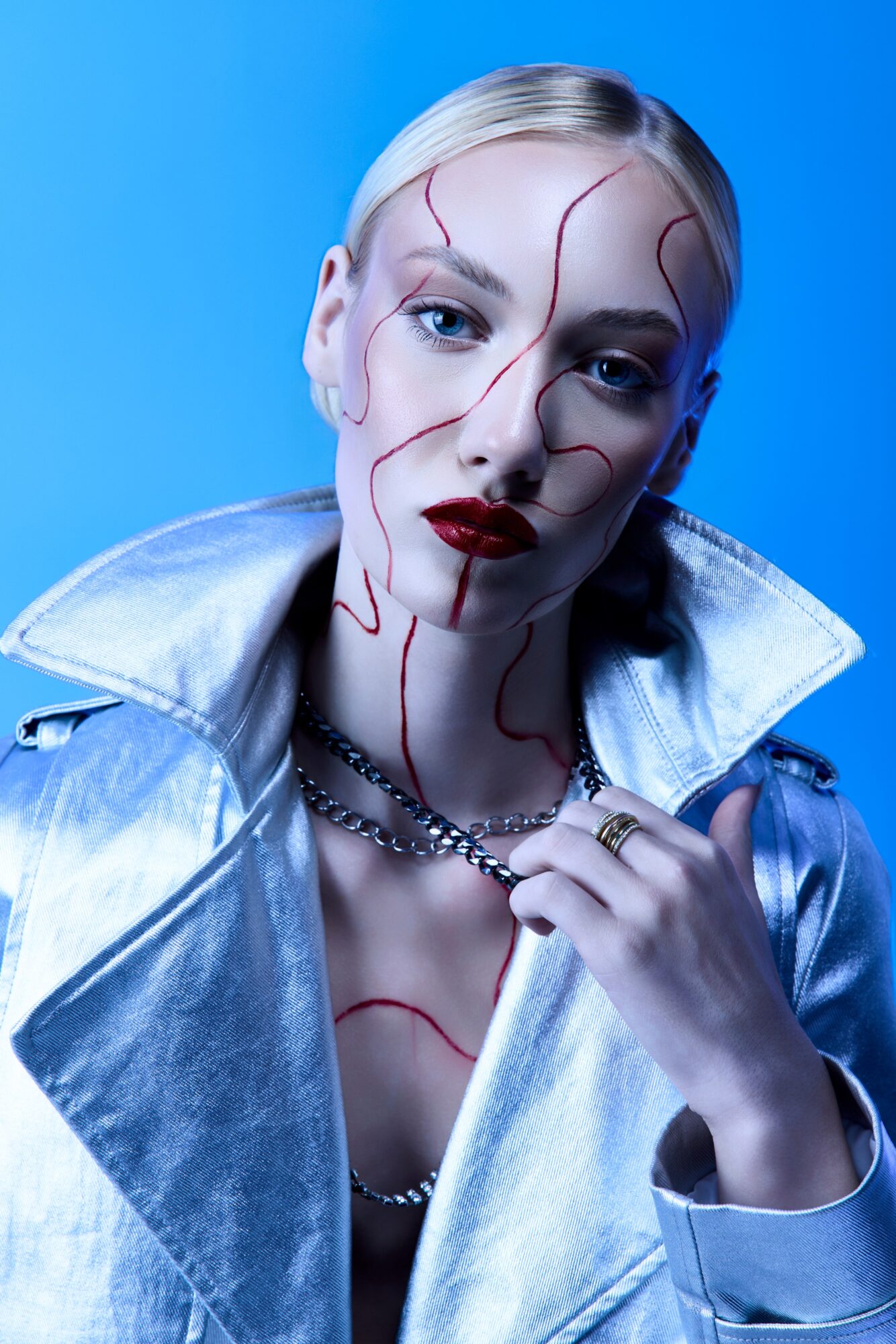 Person with pale skin and platinum blonde hair, wearing metallic jacket and jewelry, with red lines on face against blue background.
