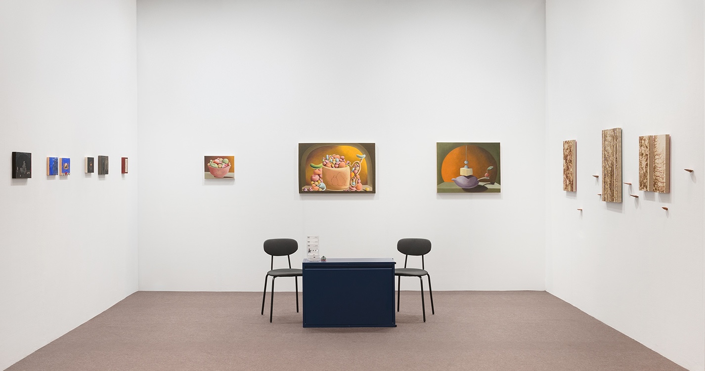 Gallery room with framed artworks on white walls and a table with two chairs.