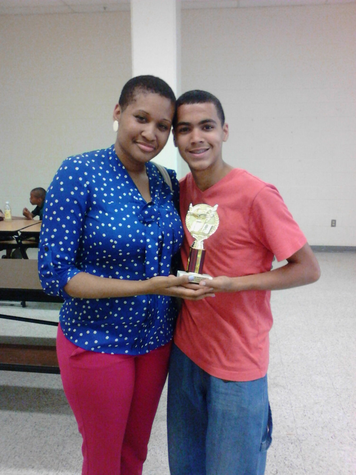 Two people stand together holding a trophy in a room with tables and benches.