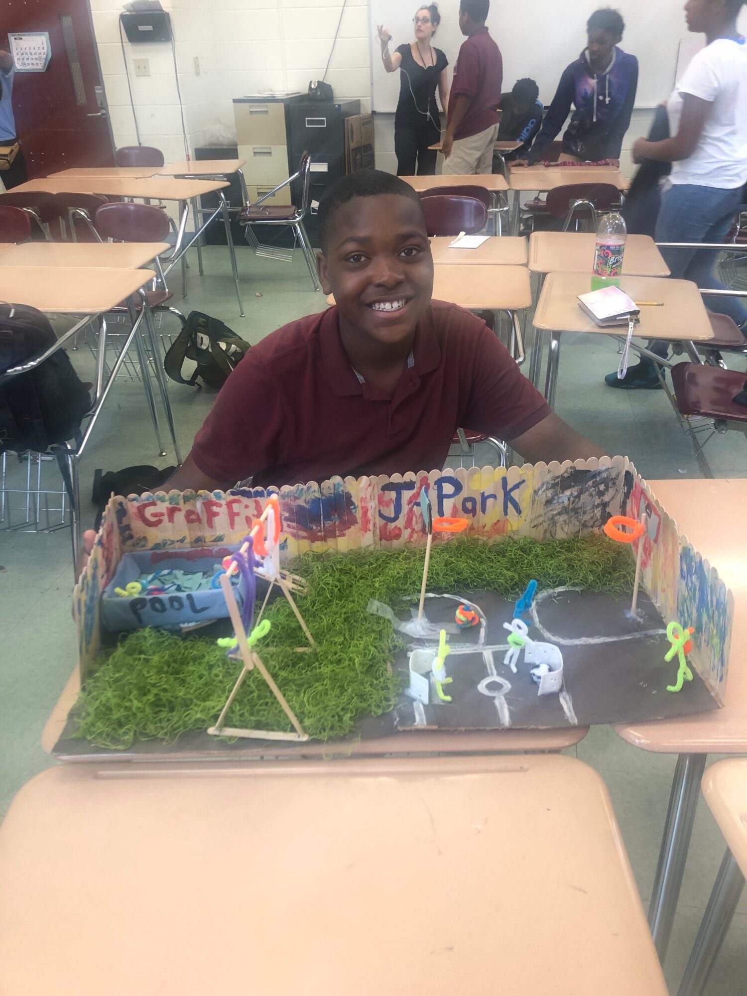 Person seated behind a classroom diorama on a desk, with students and rows of tables in the background.