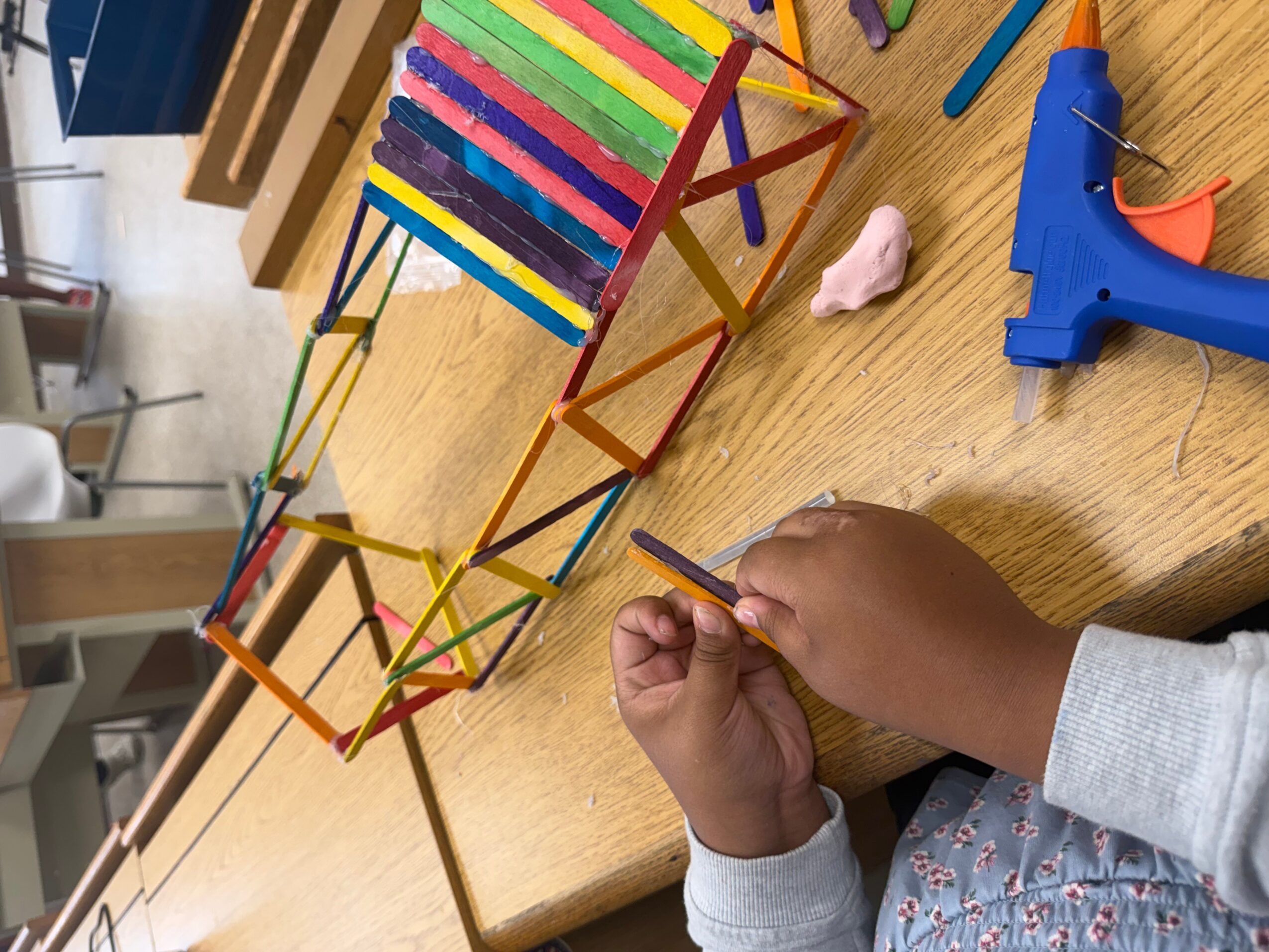 Hands assemble a colorful craft-stick structure on a wooden table beside a blue glue gun.
