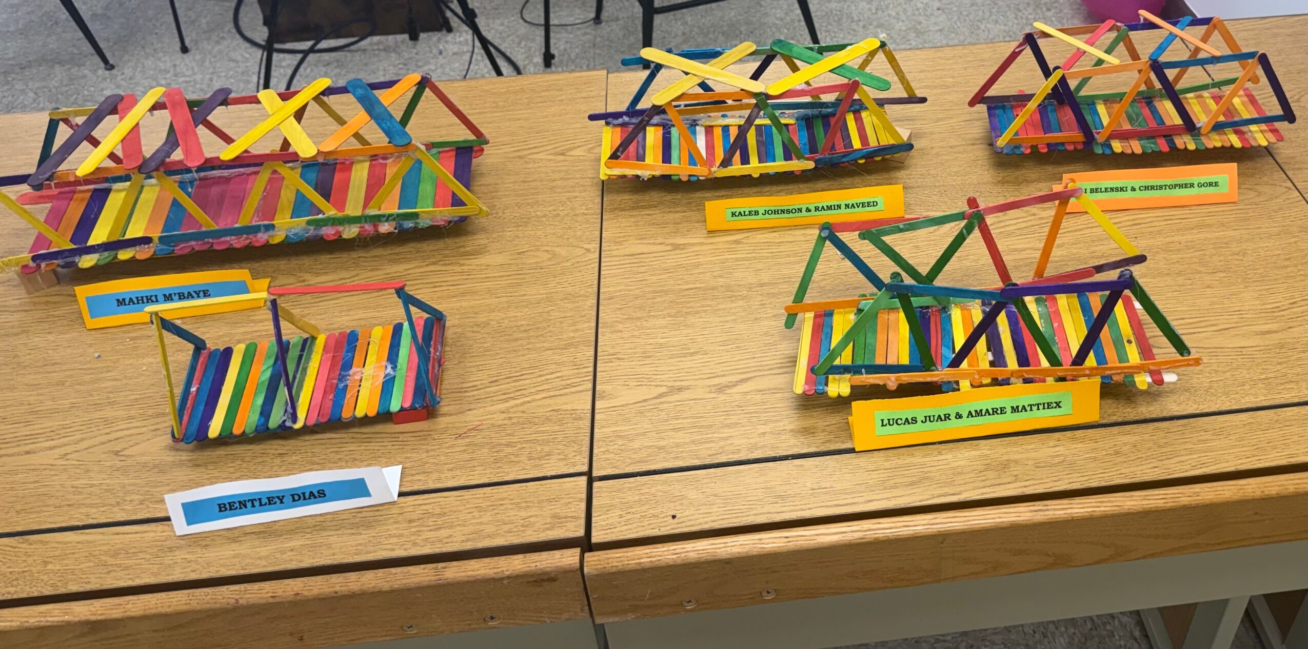 Colorful craft-stick bridges displayed on a wooden table with labels.