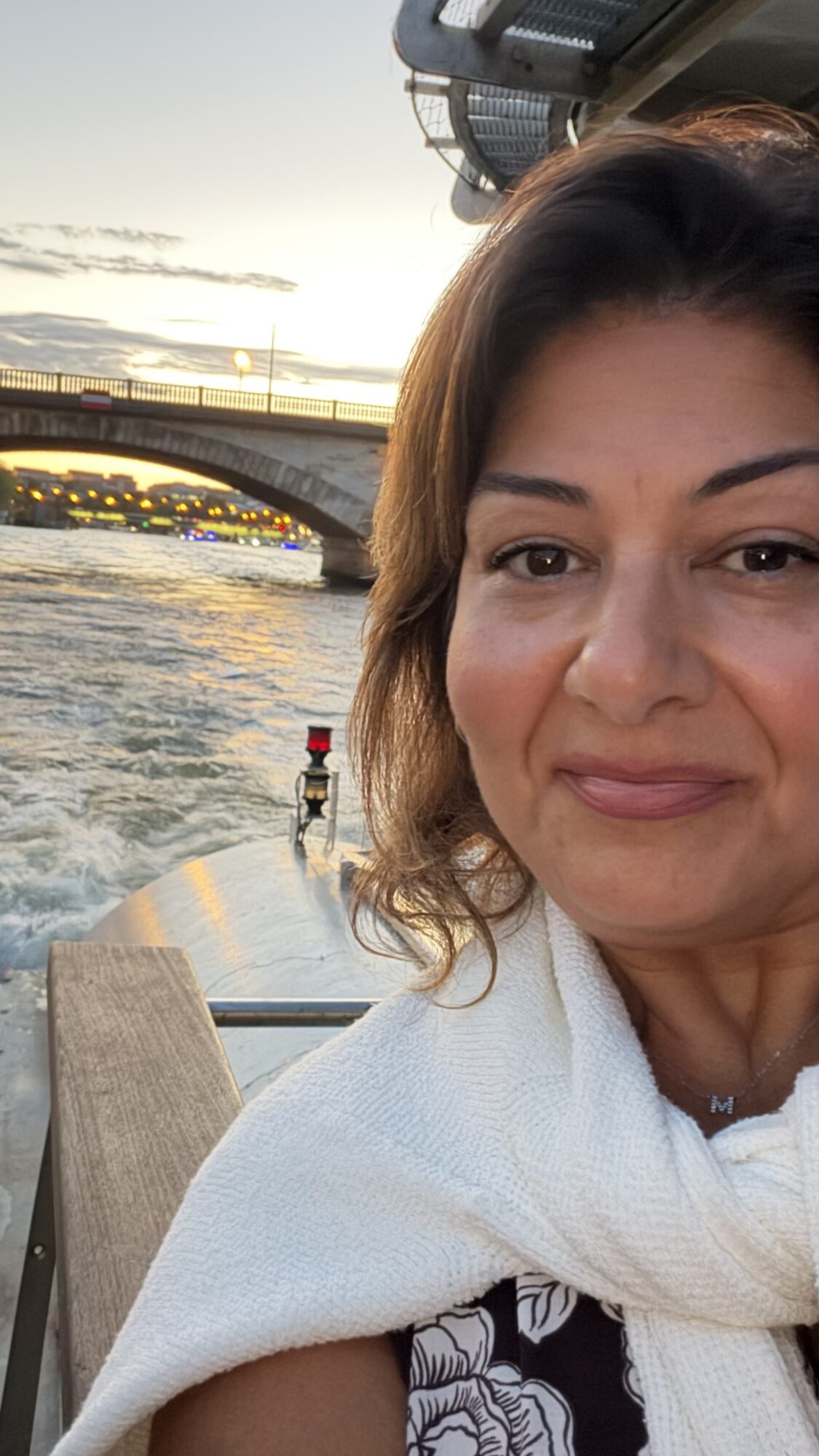 A person wrapped in a white towel sits beside choppy water, with a bridge and sunset sky behind them.