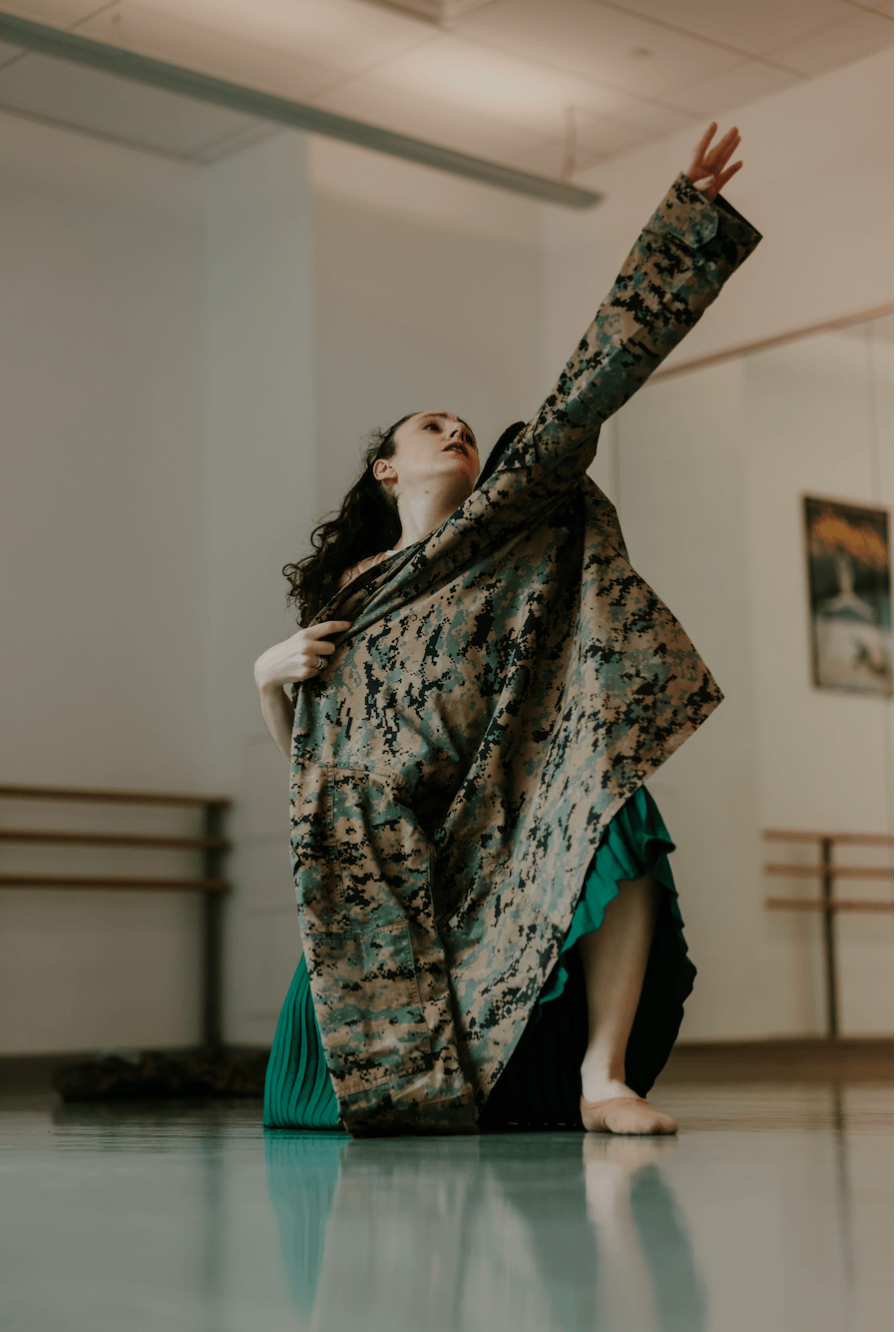Person in a dance pose with one arm raised, wearing a patterned coat and green skirt, in a dance studio.