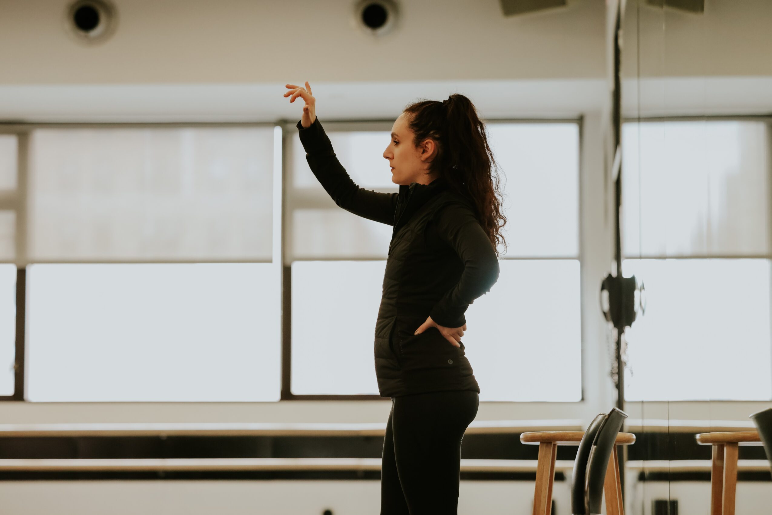 Side view of a woman in black workout clothes stretching in a gym with large windows behind her.
