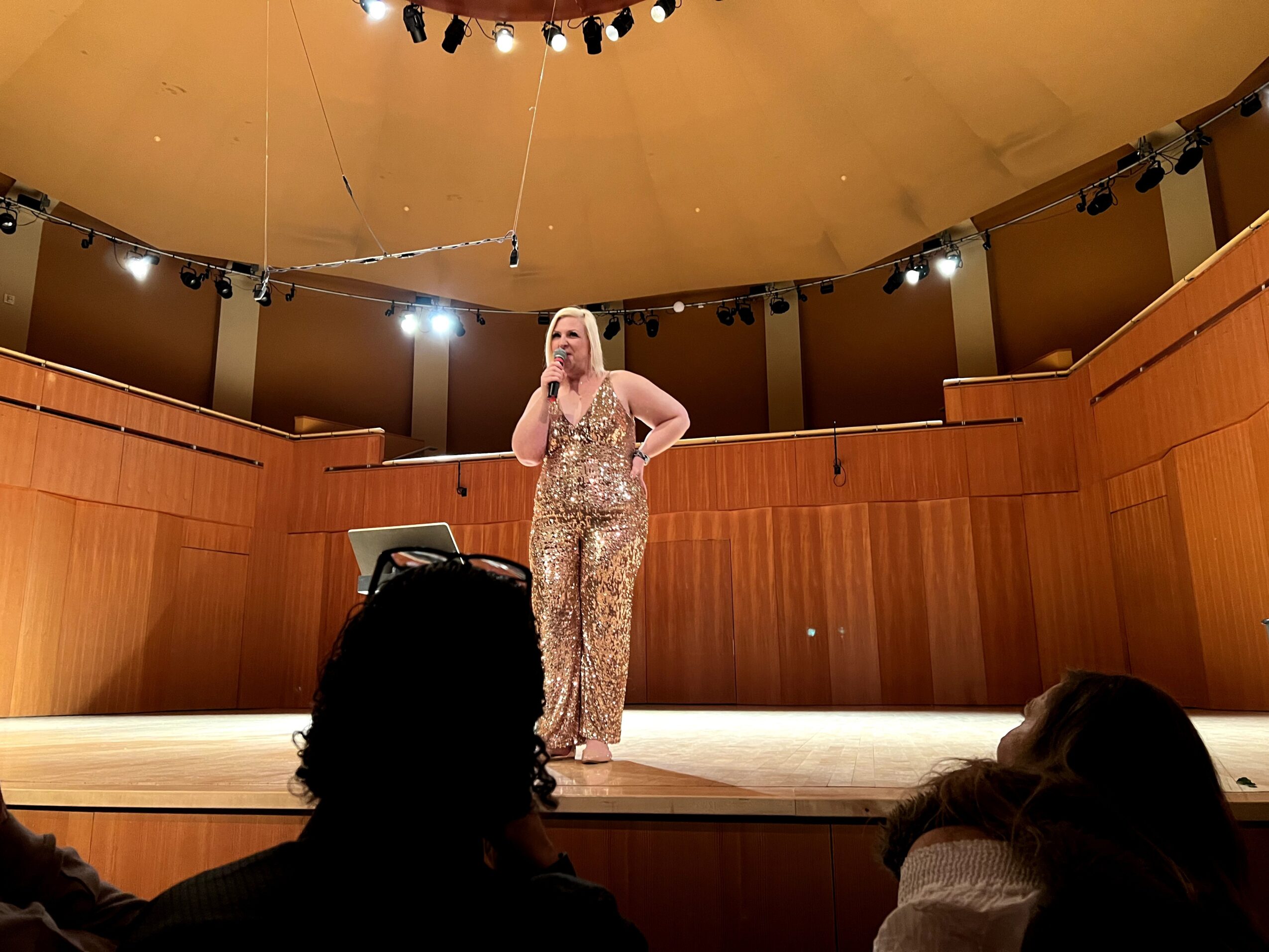 A woman in a sparkly outfit stands on stage holding a microphone, with a wooden backdrop and stage lighting.