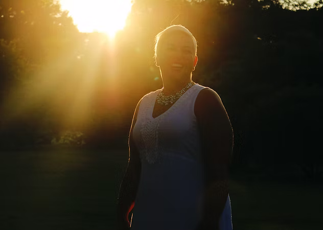 Person standing outdoors with sunlight behind, wearing a sleeveless dress, trees in background.
