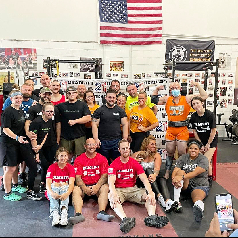 A group poses for a photo in a gym beneath an American flag, surrounded by weightlifting equipment and posters.