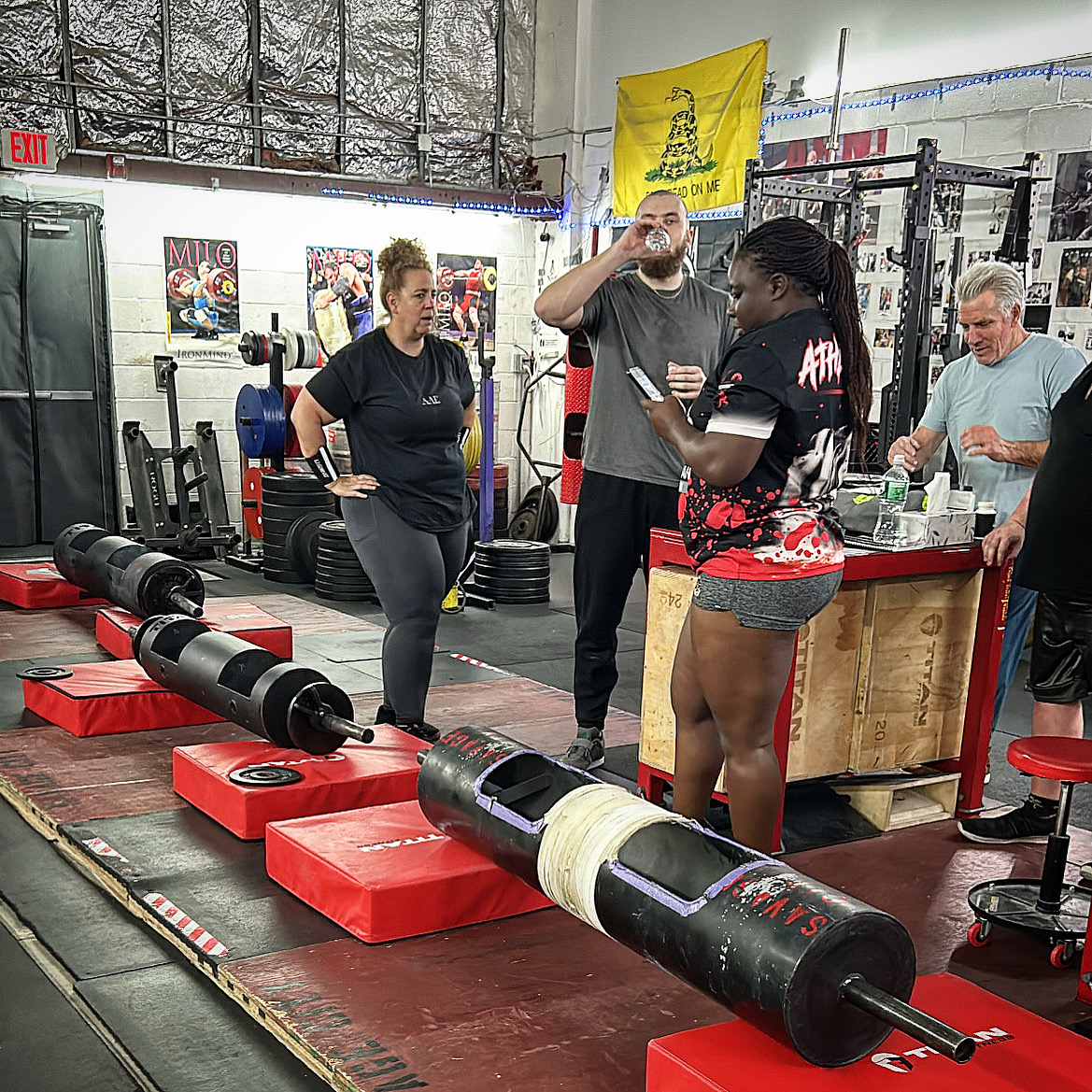 People stand around a large cylindrical metal apparatus in a gym, with red platforms and equipment in the background.