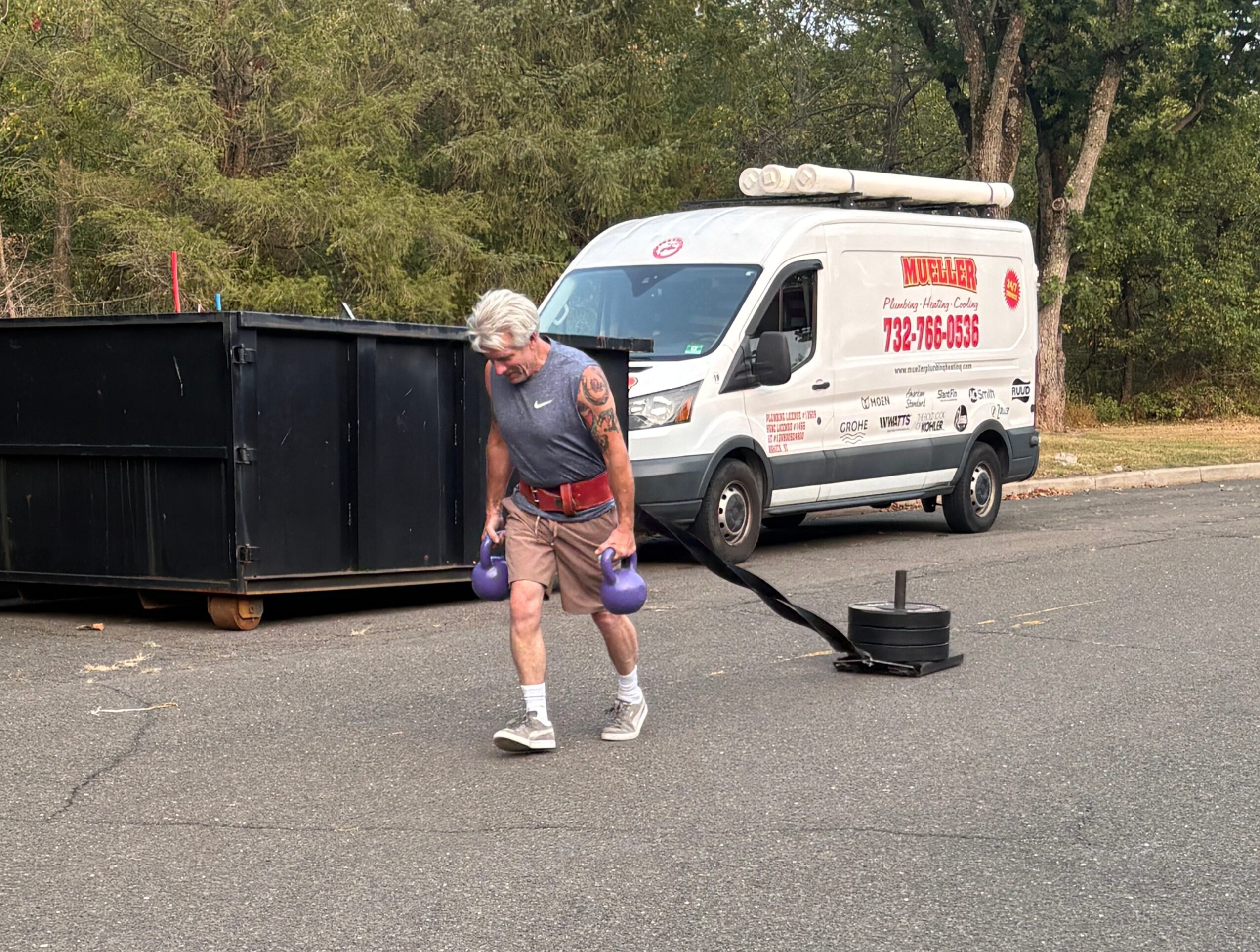 A person walks past a large black container while pulling a small wheeled cart; a white service van is parked nearby.