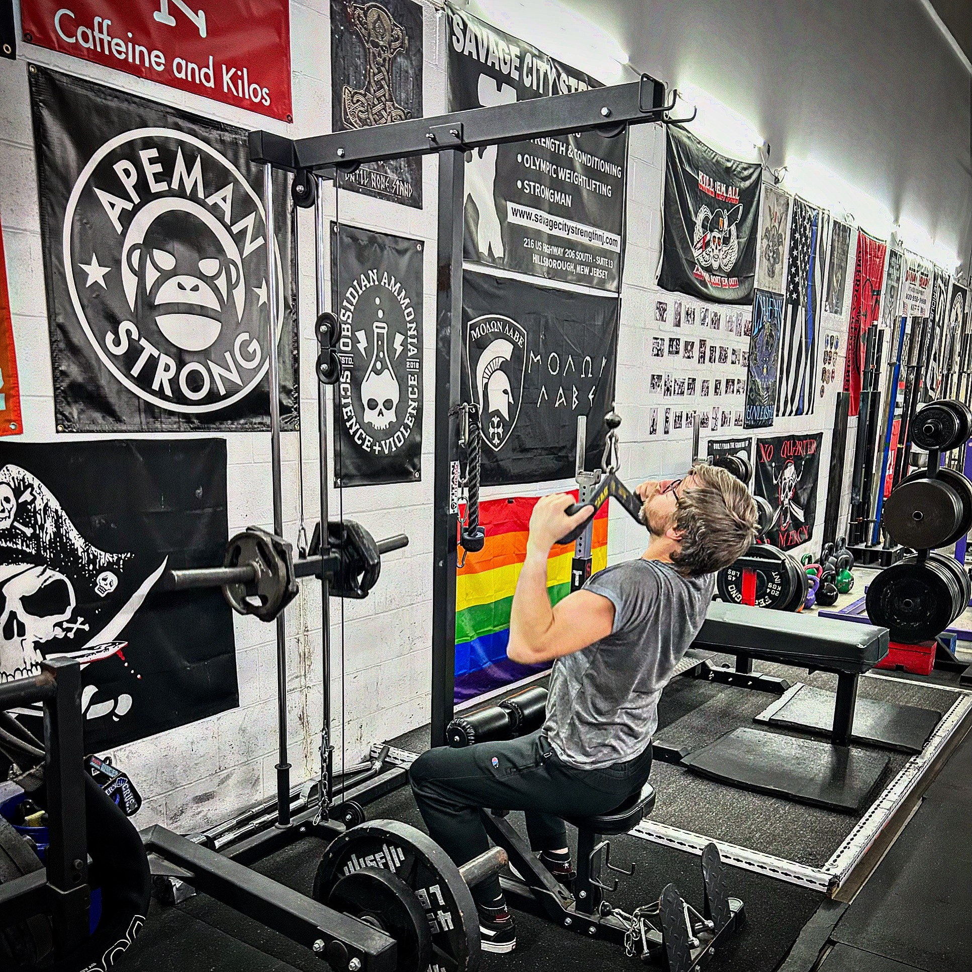 Person seated at a gym rack lifting a barbell, with weight plates and banners on the walls.