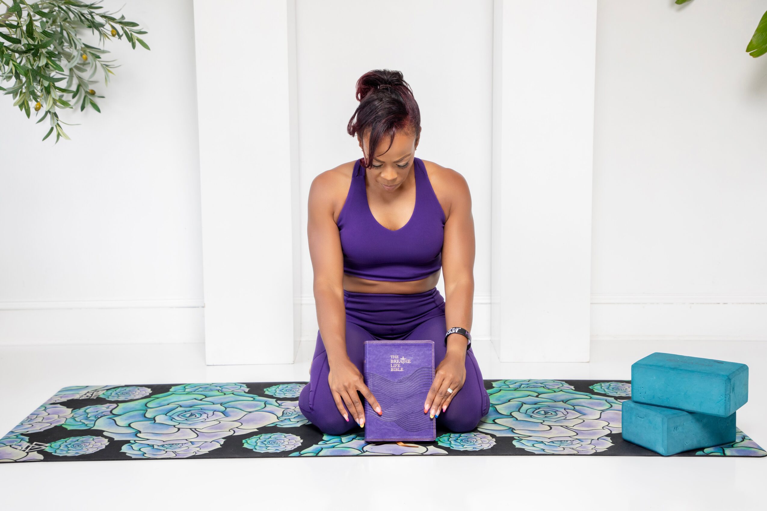 Person sitting on a yoga mat, holding a purple book, in a bright room with plants and blue blocks nearby.