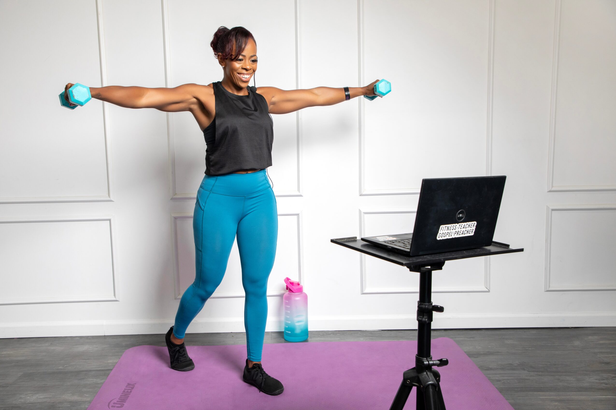 Woman in workout clothes holding dumbbells with arms extended, standing on a purple mat in front of a laptop, in a room with white walls.