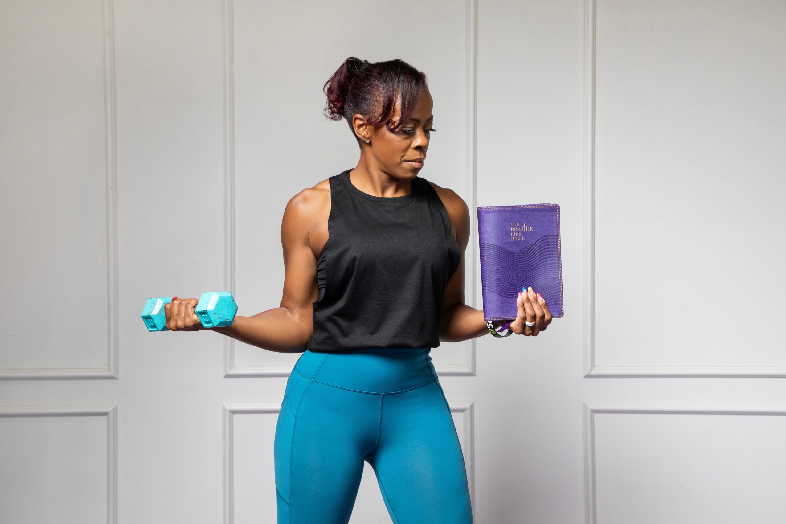 Woman in workout clothes holding a dumbbell and a tablet, standing against a plain wall.