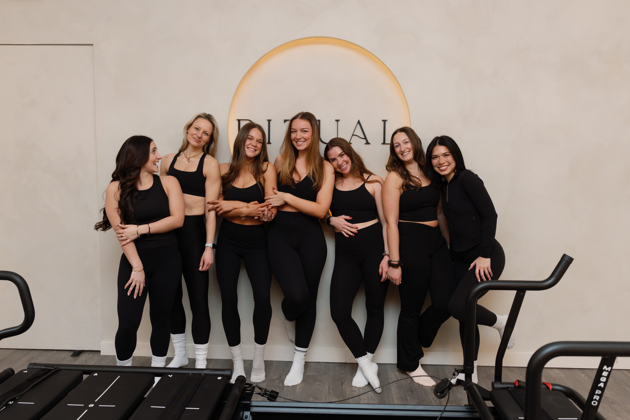Six women standing together in front of a wall with a circular logo, smiling, dressed in black outfits, some with arms around each other.
