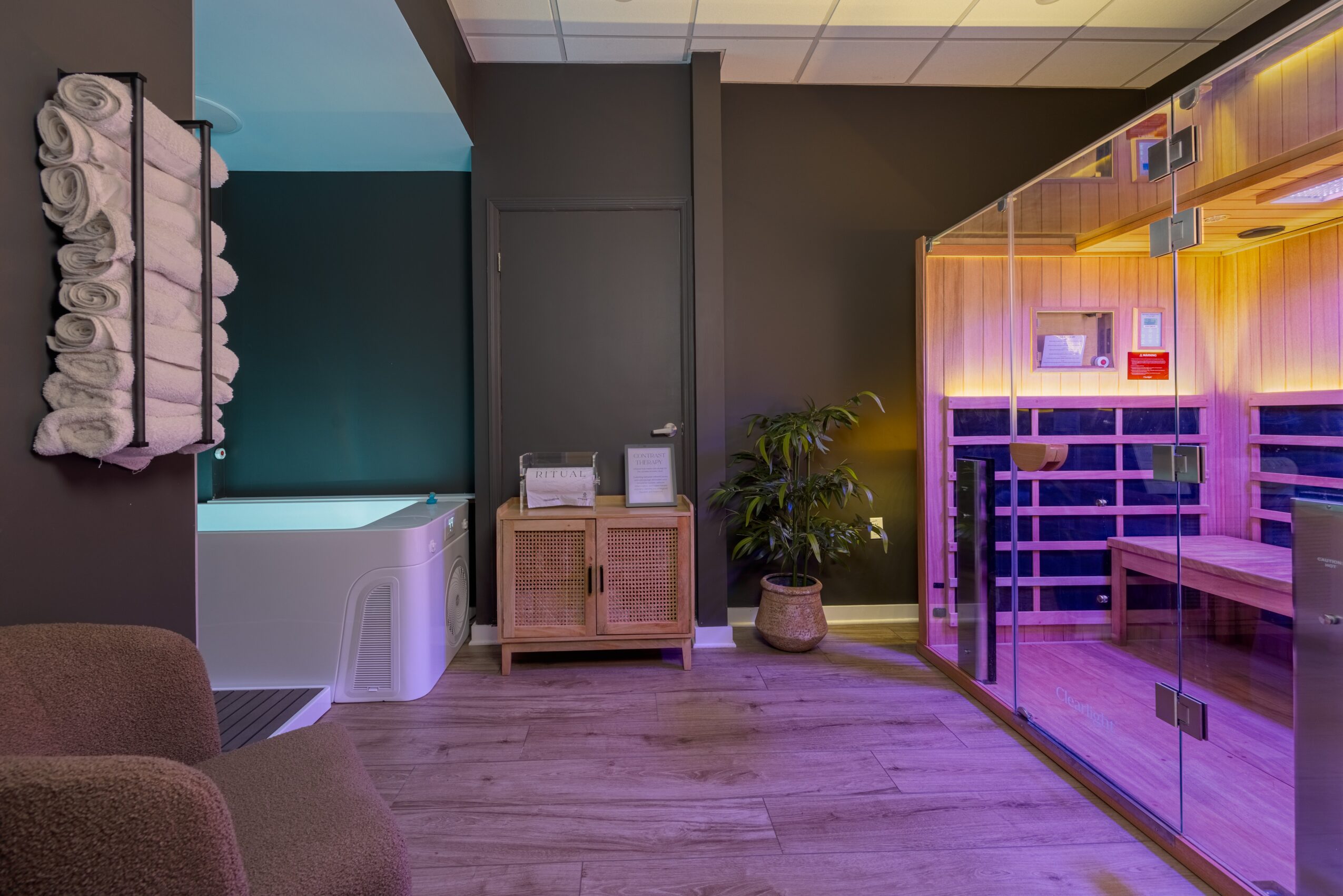 Spa room with a hot tub, towel rack, wooden cabinet, potted plant, and a glass sauna with purple lighting.