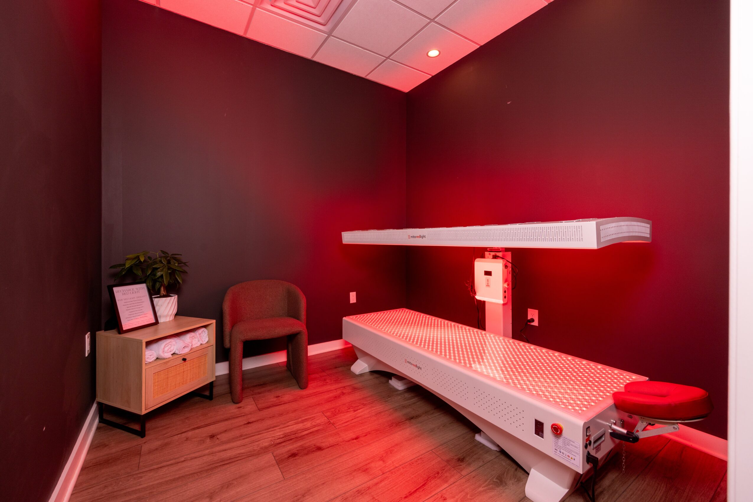 Hospital room with a bed, chair, small table, and red lighting, with dark walls and wooden floor.