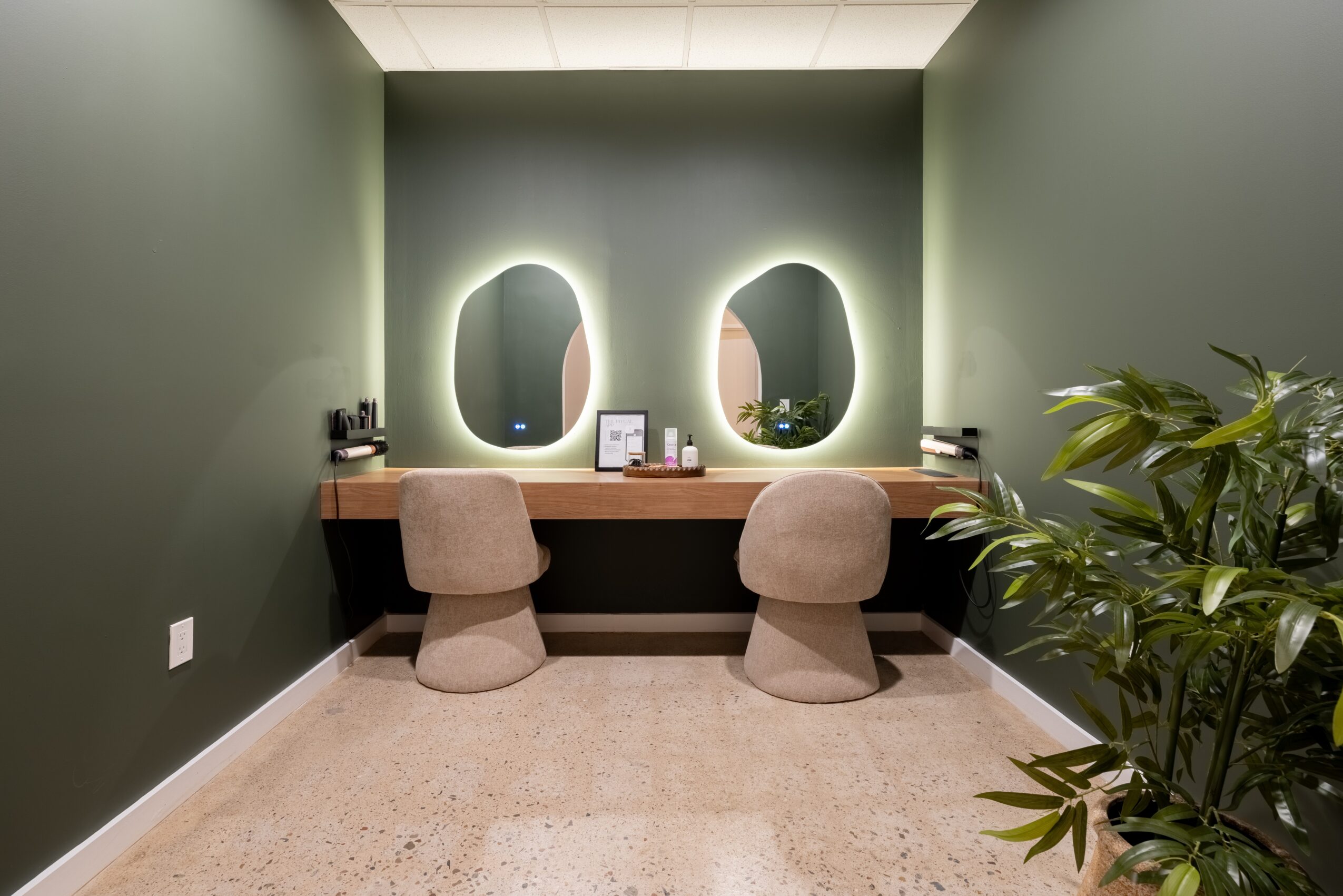 Two illuminated oval mirrors above a wooden desk with two chairs, a plant on the right, and a green wall background.