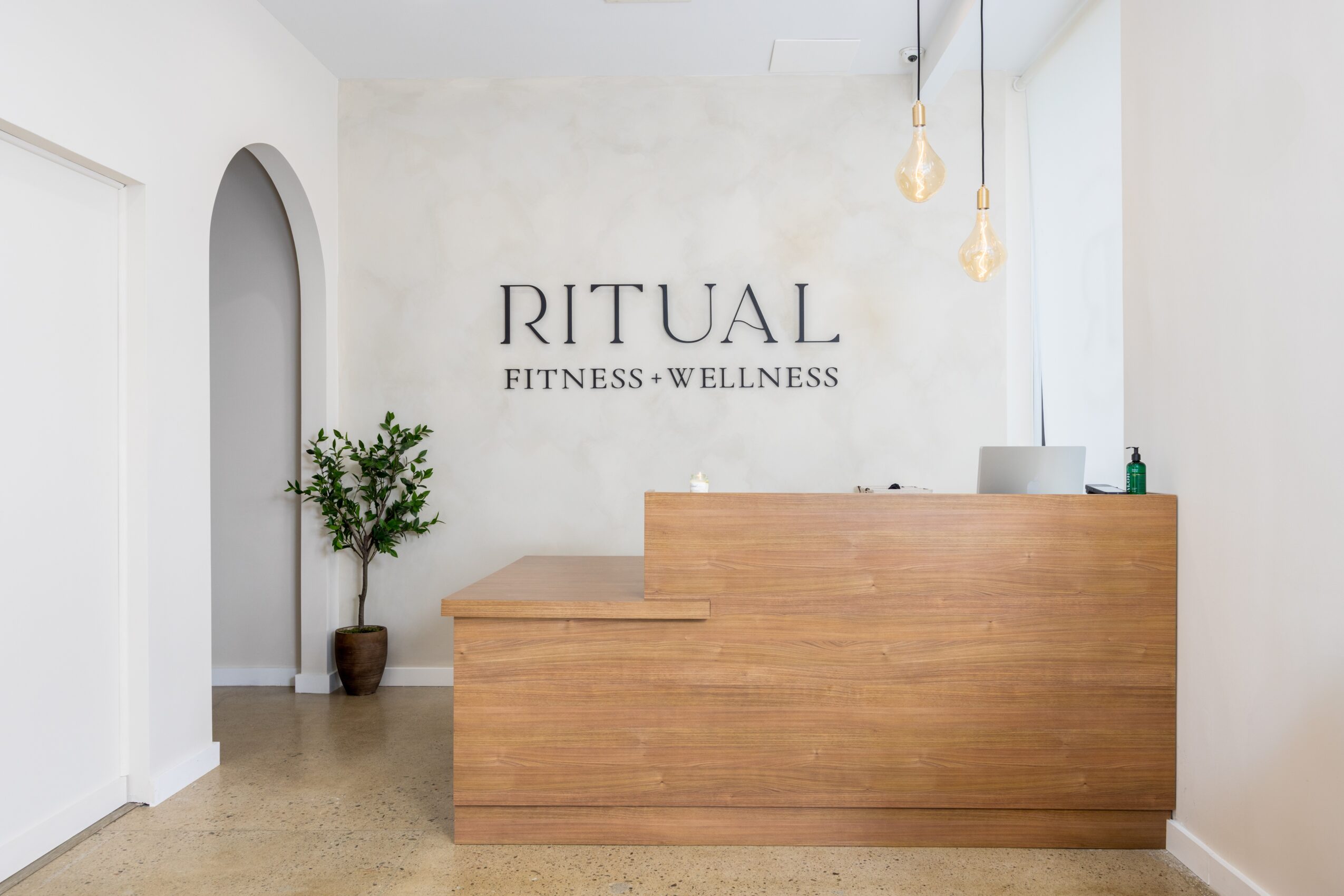 Reception desk in a wellness center with a potted plant, white walls, and hanging pendant lights, and a sign reading 'RITUAL FITNESS WELLNESS'.