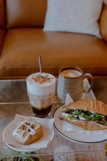 Coffee drink with whipped cream, a mug, a sandwich, a cookie, and a croissant on a glass table in front of a sofa.