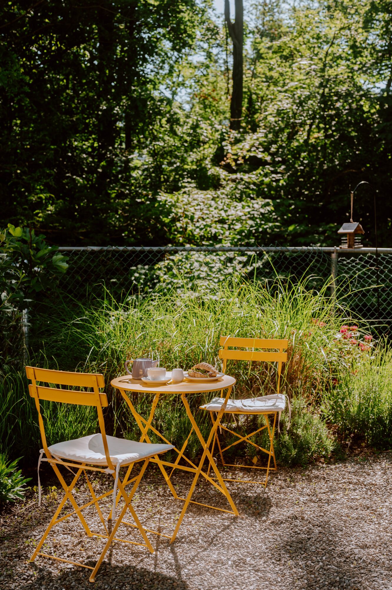 Three yellow chairs around a small table in a garden with lush greenery and a chain-link fence in the background.
