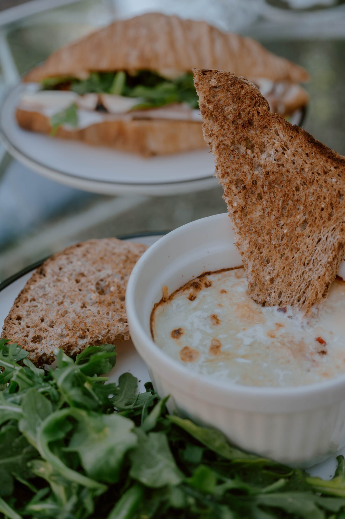 A bowl of creamy soup with a toasted bread triangle, fresh greens, and a sandwich in the background.