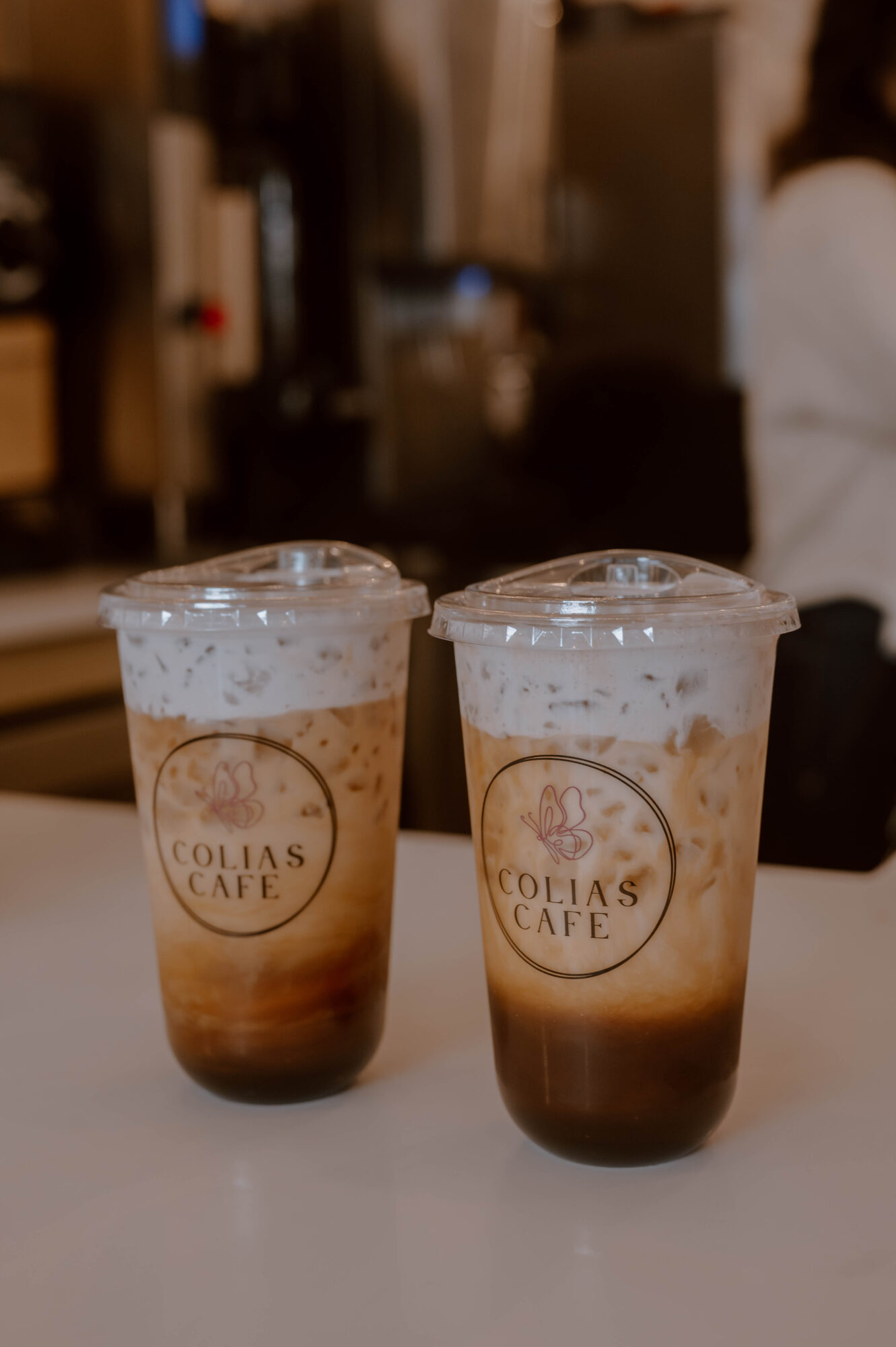Two iced coffee drinks in clear cups with lids, placed on a white surface, with a blurred background.