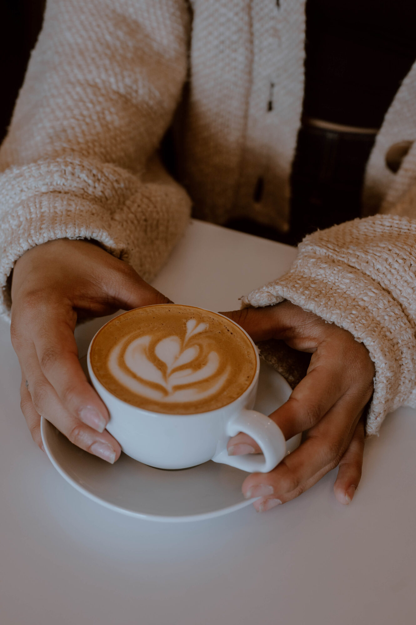 Person holding a cup of coffee with latte art, sitting at a table, wearing a beige sweater.