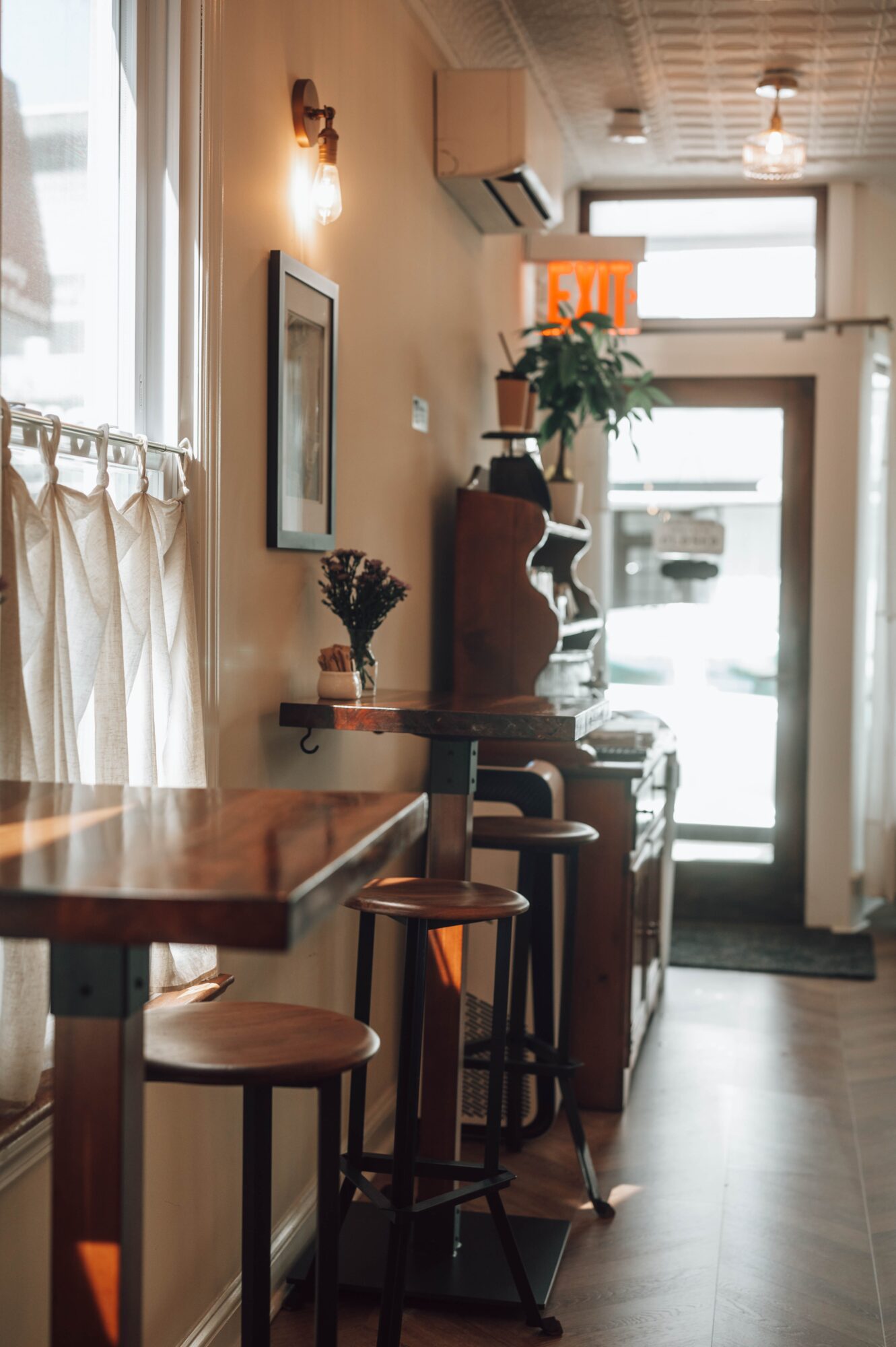 Interior of a cozy cafe with a wooden counter, bar stools, a potted plant, and a window with curtains.