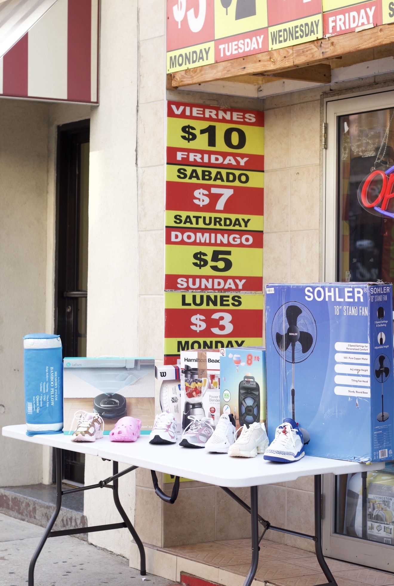 Table with shoes, a blue water bottle, and a blue box outside a store, with a sign showing daily prices.