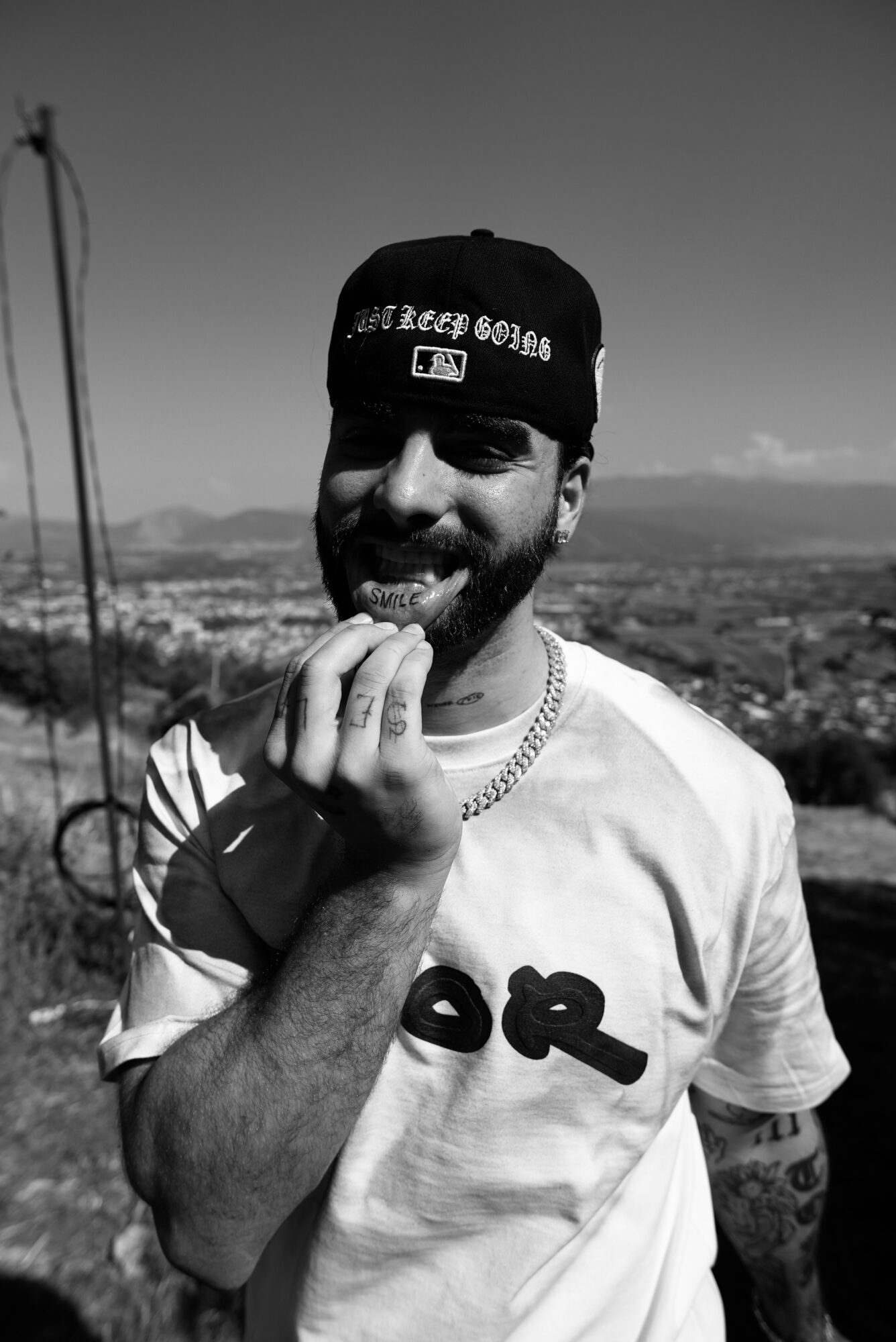 A bearded person wearing a backward cap and T-shirt stands outdoors, holding a small object near their mouth with a landscape behind them.