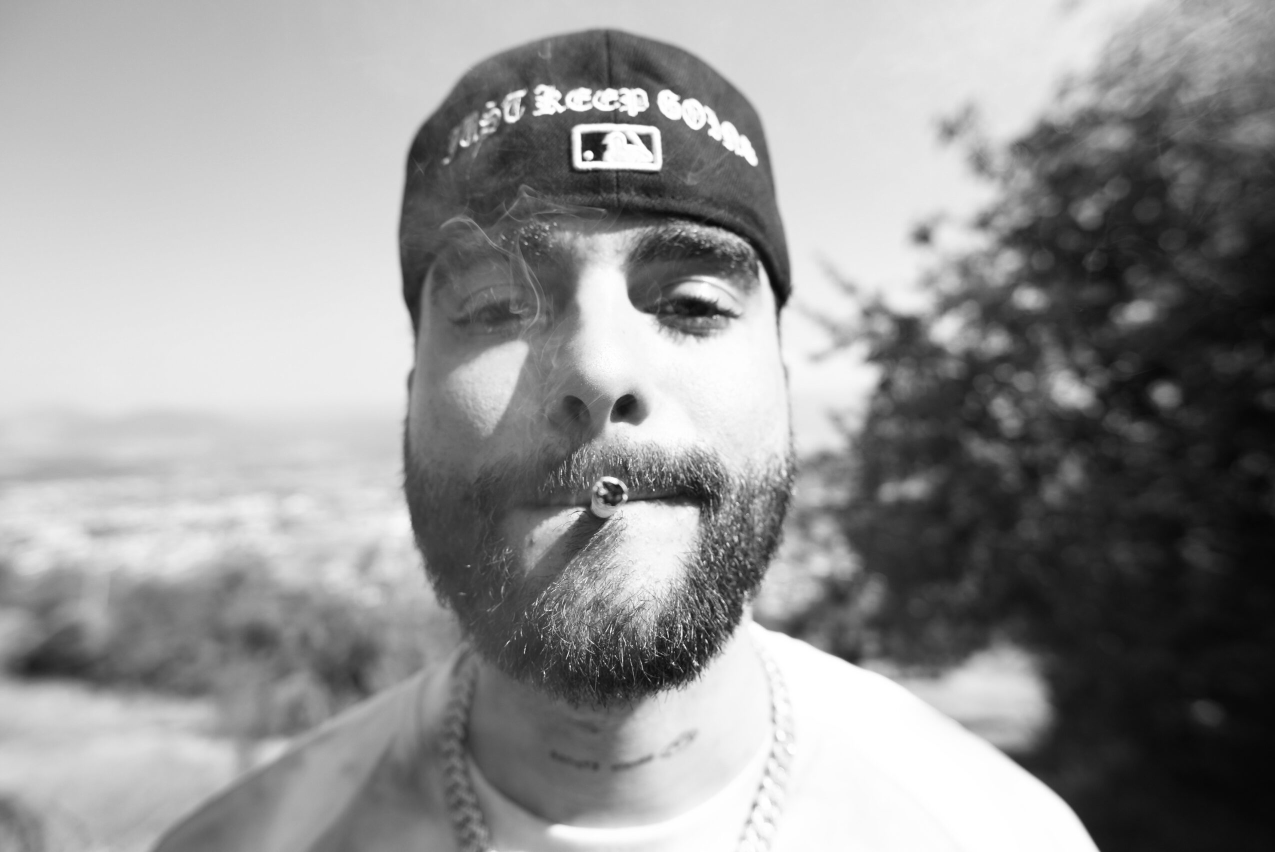 Black-and-white close-up of a bearded person wearing a backward cap, with trees and a landscape in the background.