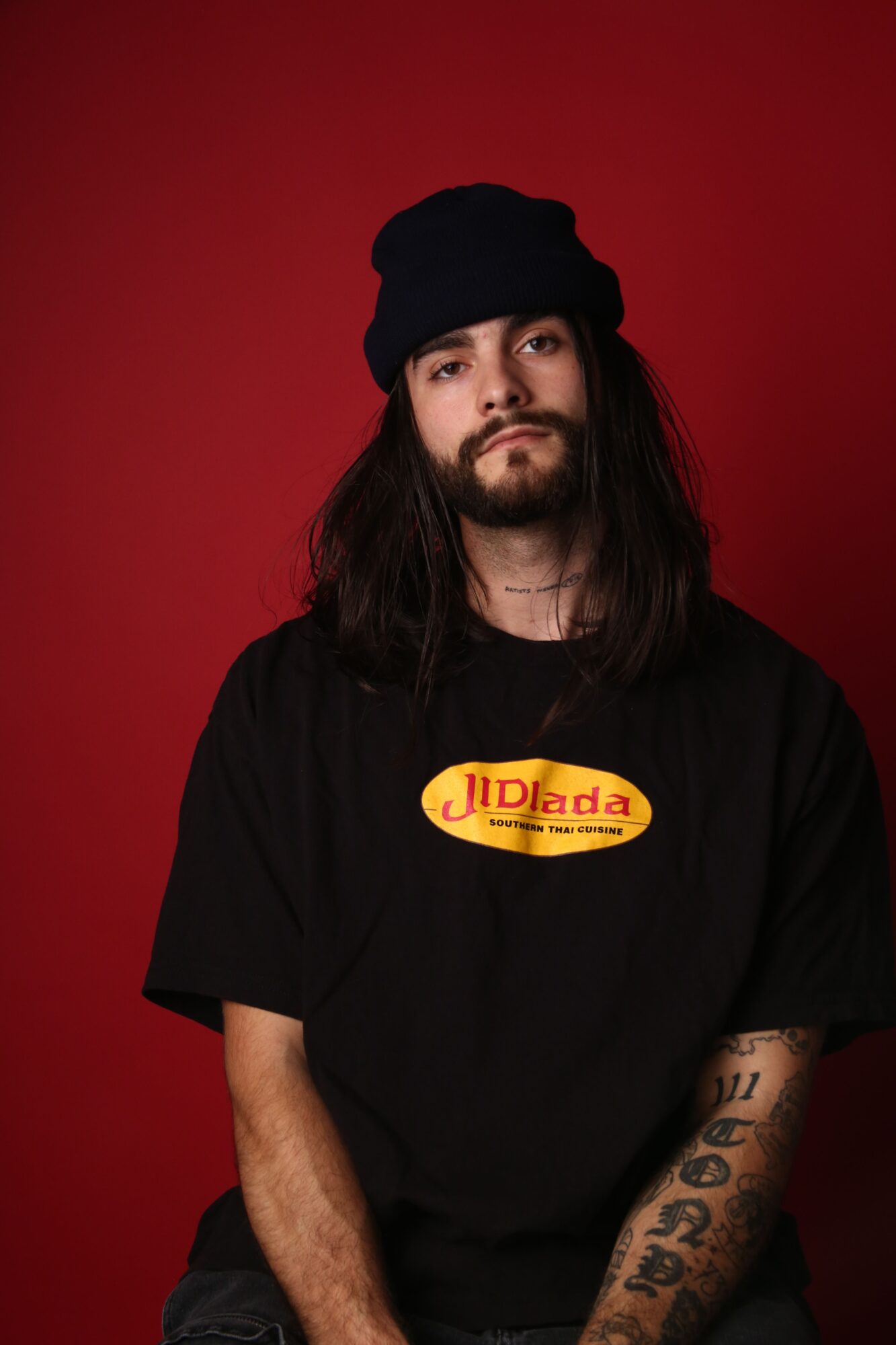 A person wearing a black cap and black shirt with a yellow-and-red logo stands against a red background.
