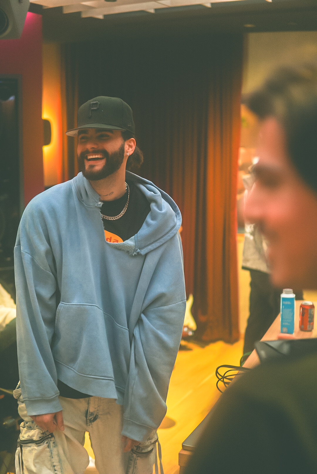 A person wearing a dark cap and blue hoodie stands beside a blurred person in the foreground.