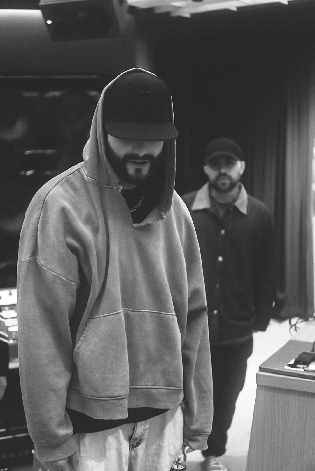 Two people in a recording studio, with one wearing a hooded sweatshirt and cap in the foreground.