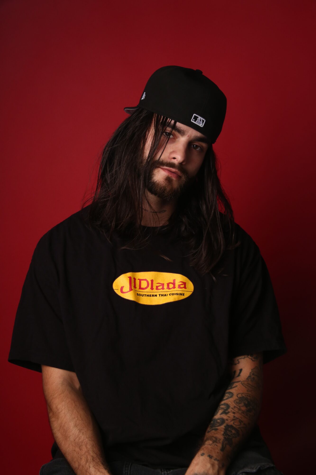 Person wearing a black cap and black T-shirt with a yellow-and-red logo against a red background.