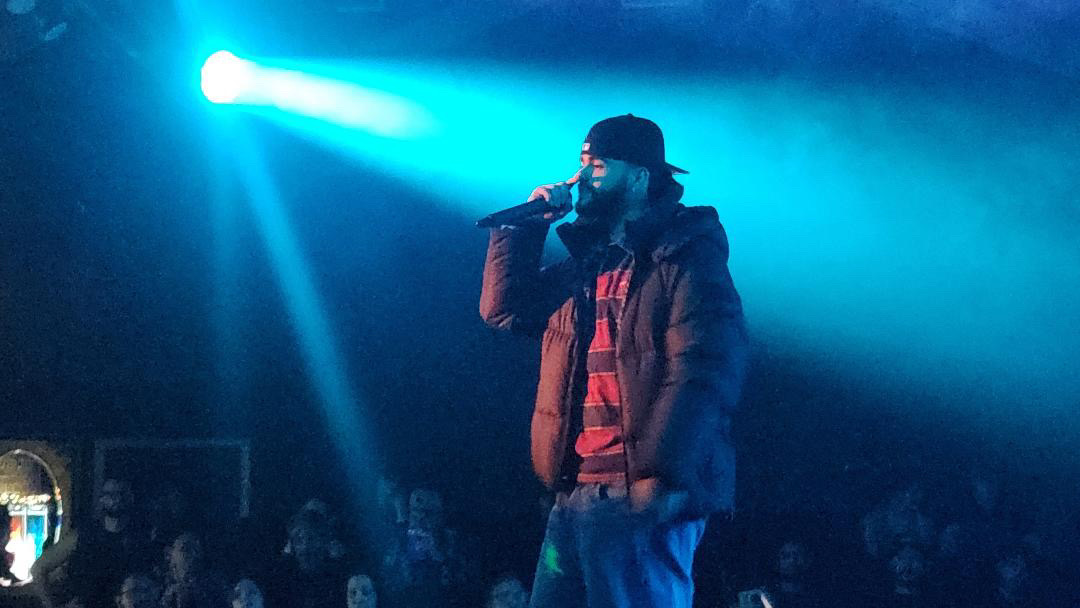 A performer holds a microphone on a dark stage under bright blue lights, with an audience below.