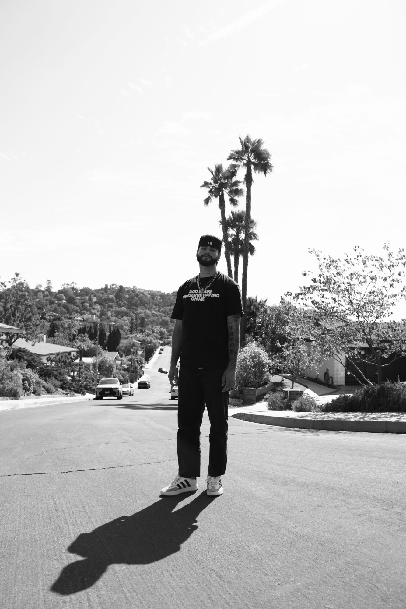 Person standing in the middle of a road, with palm trees, houses, and a long shadow.