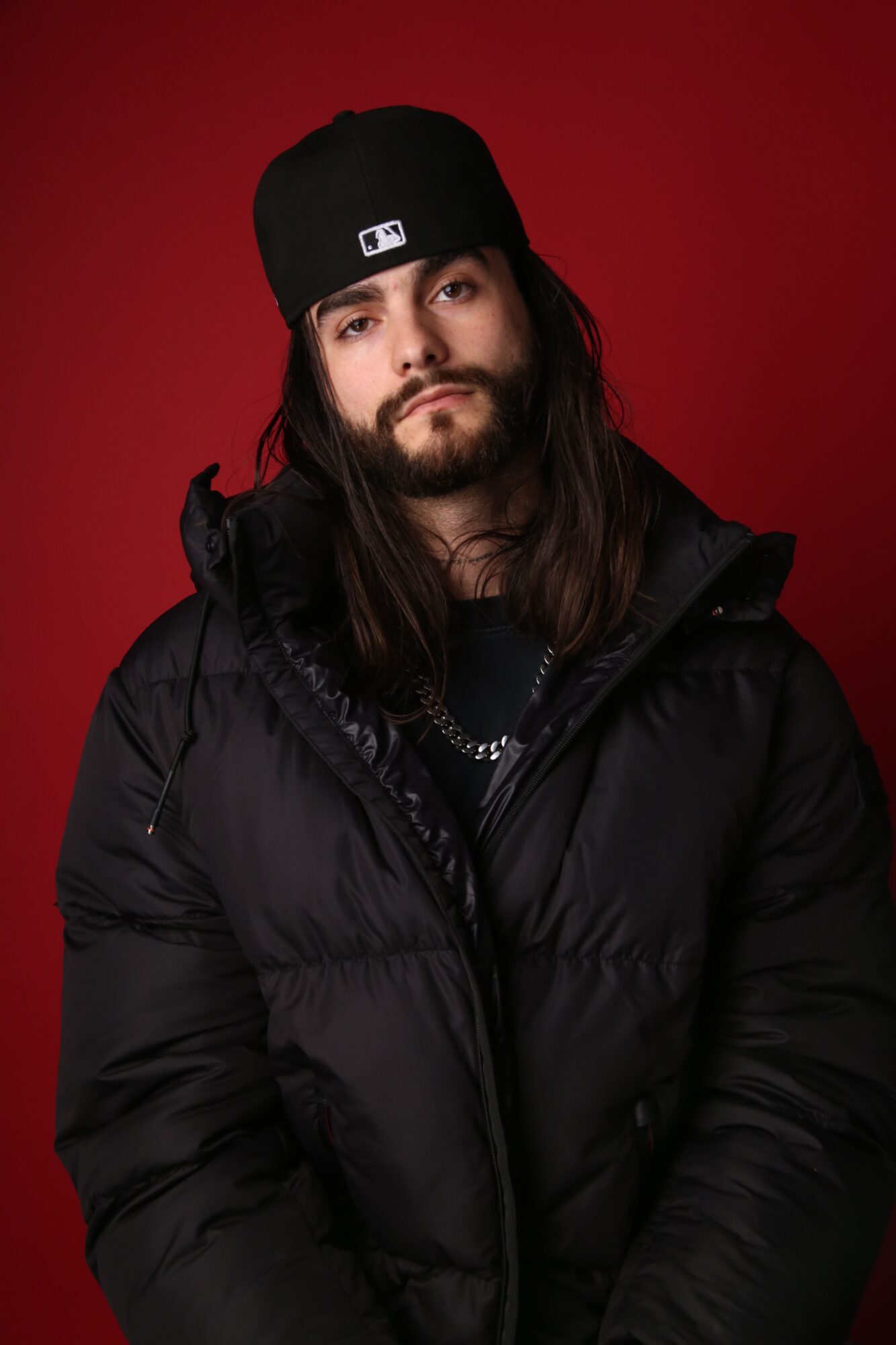 Person with long hair wearing a black cap and puffy jacket against a red background.