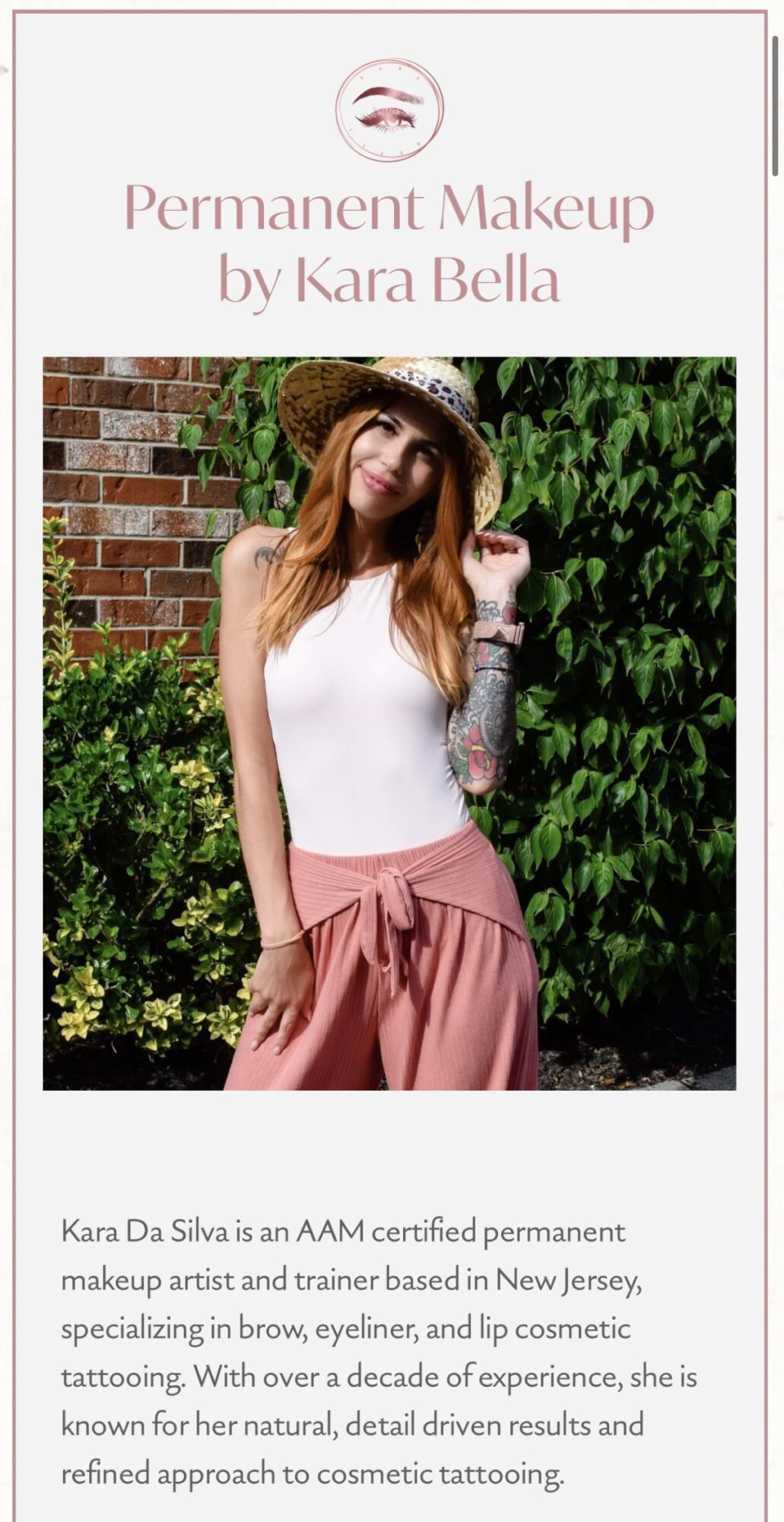 A woman with long hair wearing a hat, white top, and pink pants standing outdoors with greenery and brick wall behind her.