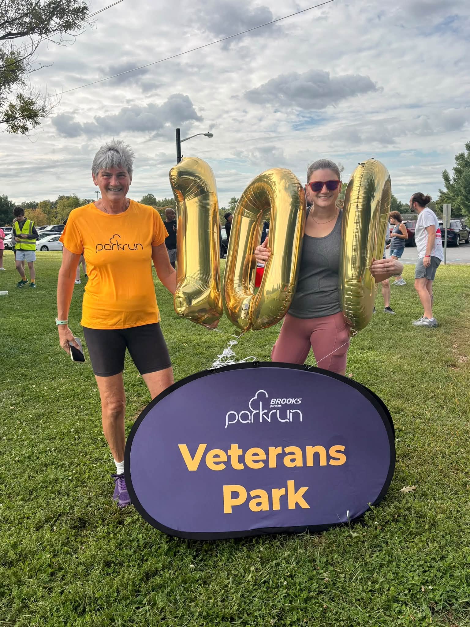 Two people stand beside gold number balloons and a Veterans Park sign on a grassy field.