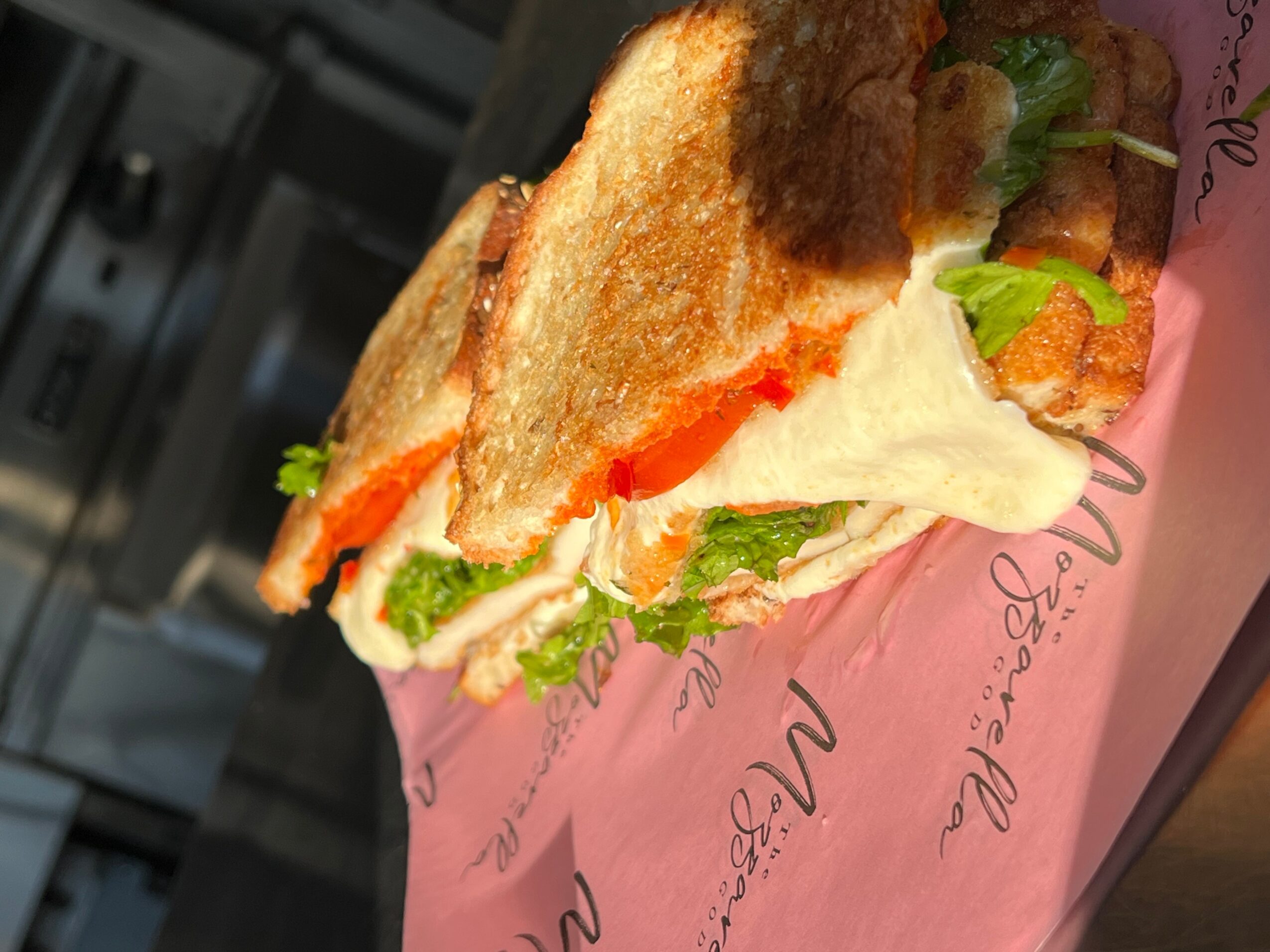 Sandwich with toasted bread, lettuce, tomato, cheese, and meat, wrapped in pink paper.