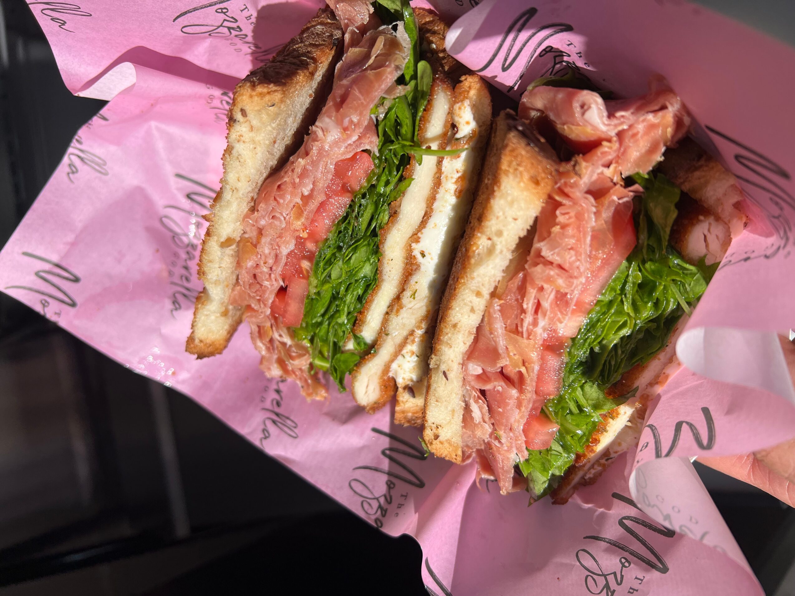 Close-up of a sandwich with bread, lettuce, tomato, and meat, wrapped in pink paper.