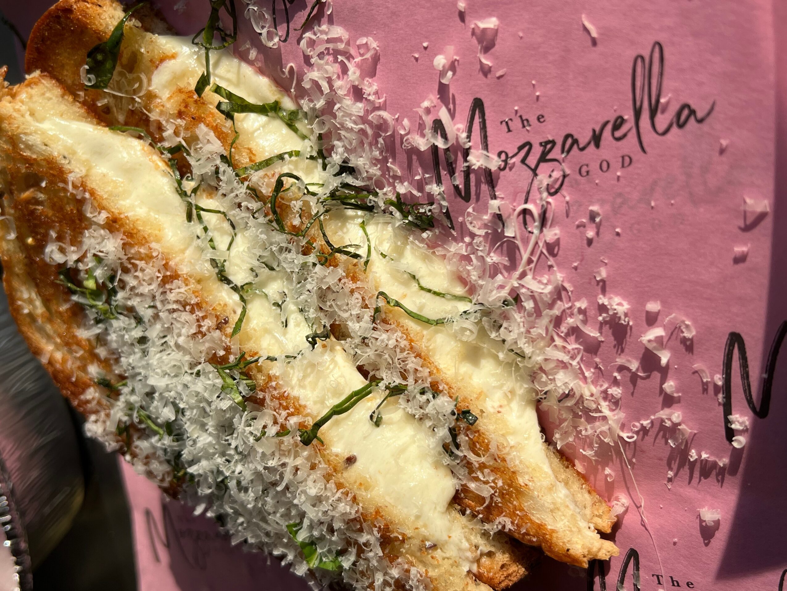 Breaded mozzarella stick with herbs and grated cheese on pink paper, with text reading 'Mozzarella' and 'God'.