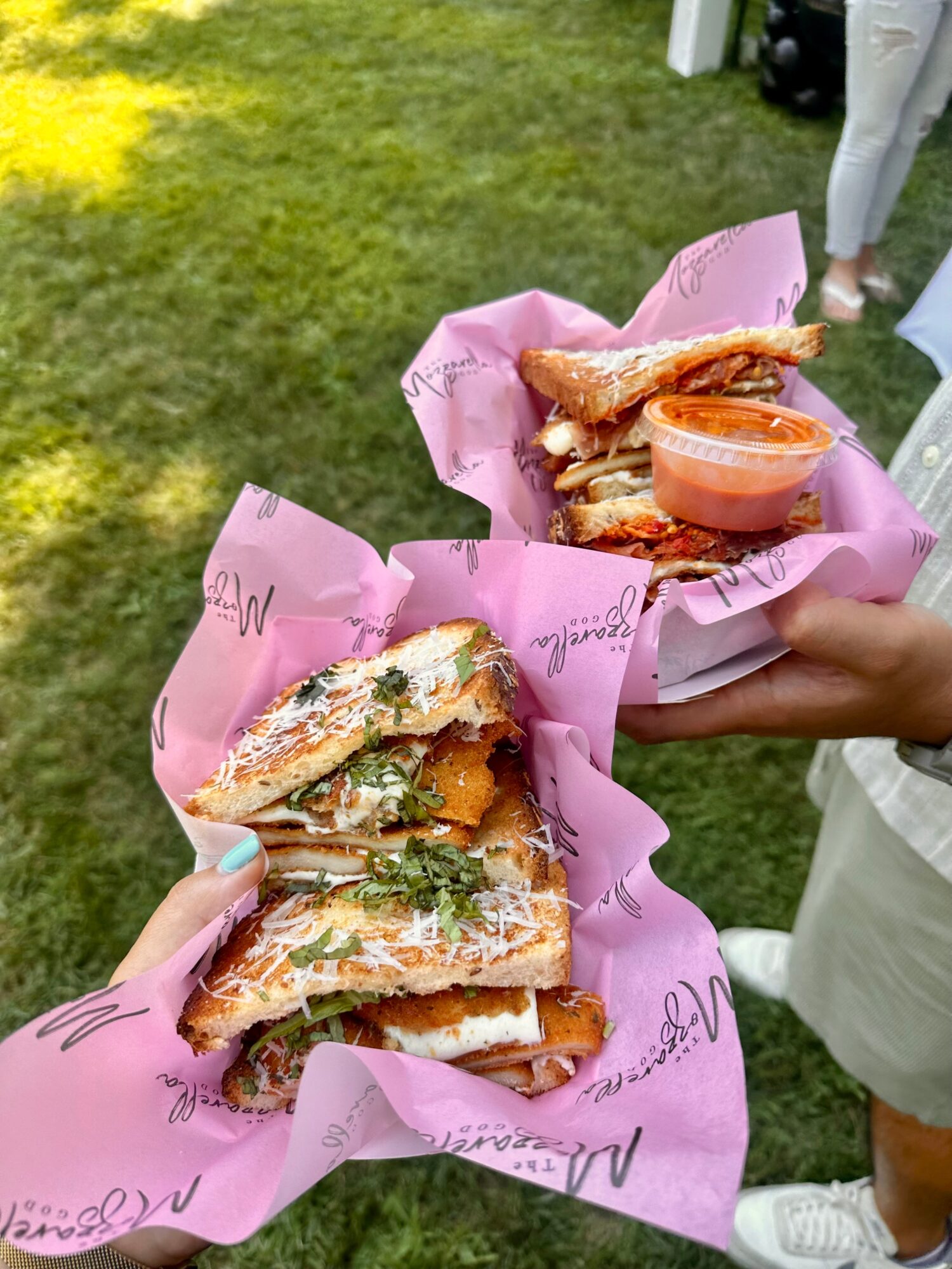 Two hands hold sandwiches wrapped in pink paper, with a side of sauce, outdoors on grass.
