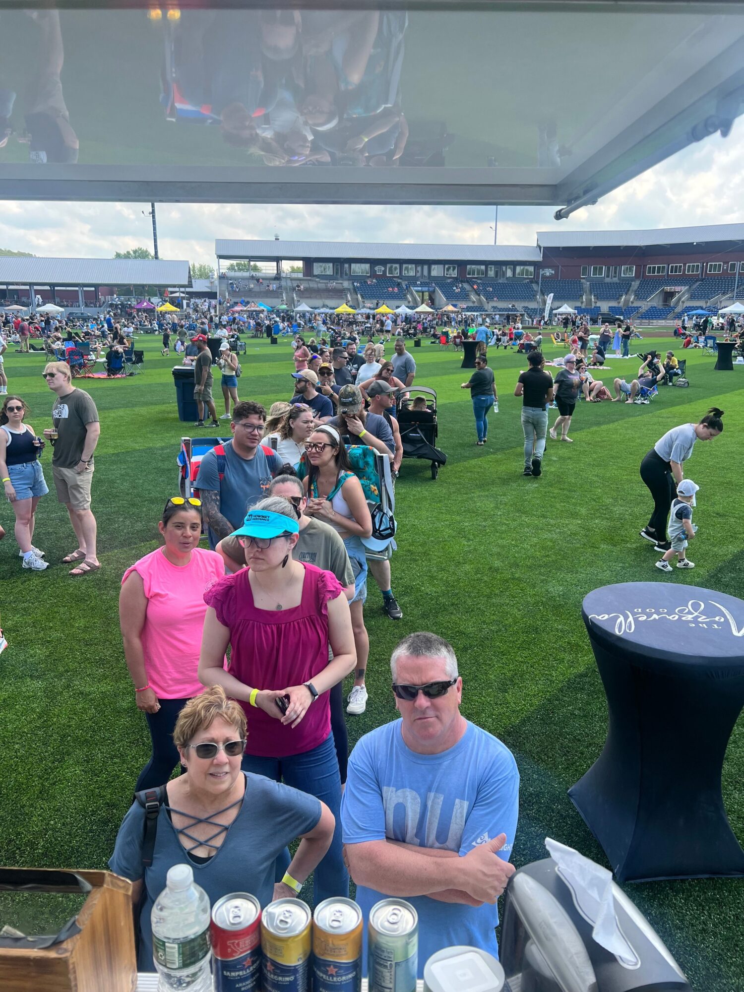 People gathered outdoors on a grassy area, some standing and others sitting, with a large screen and event setup in the background.
