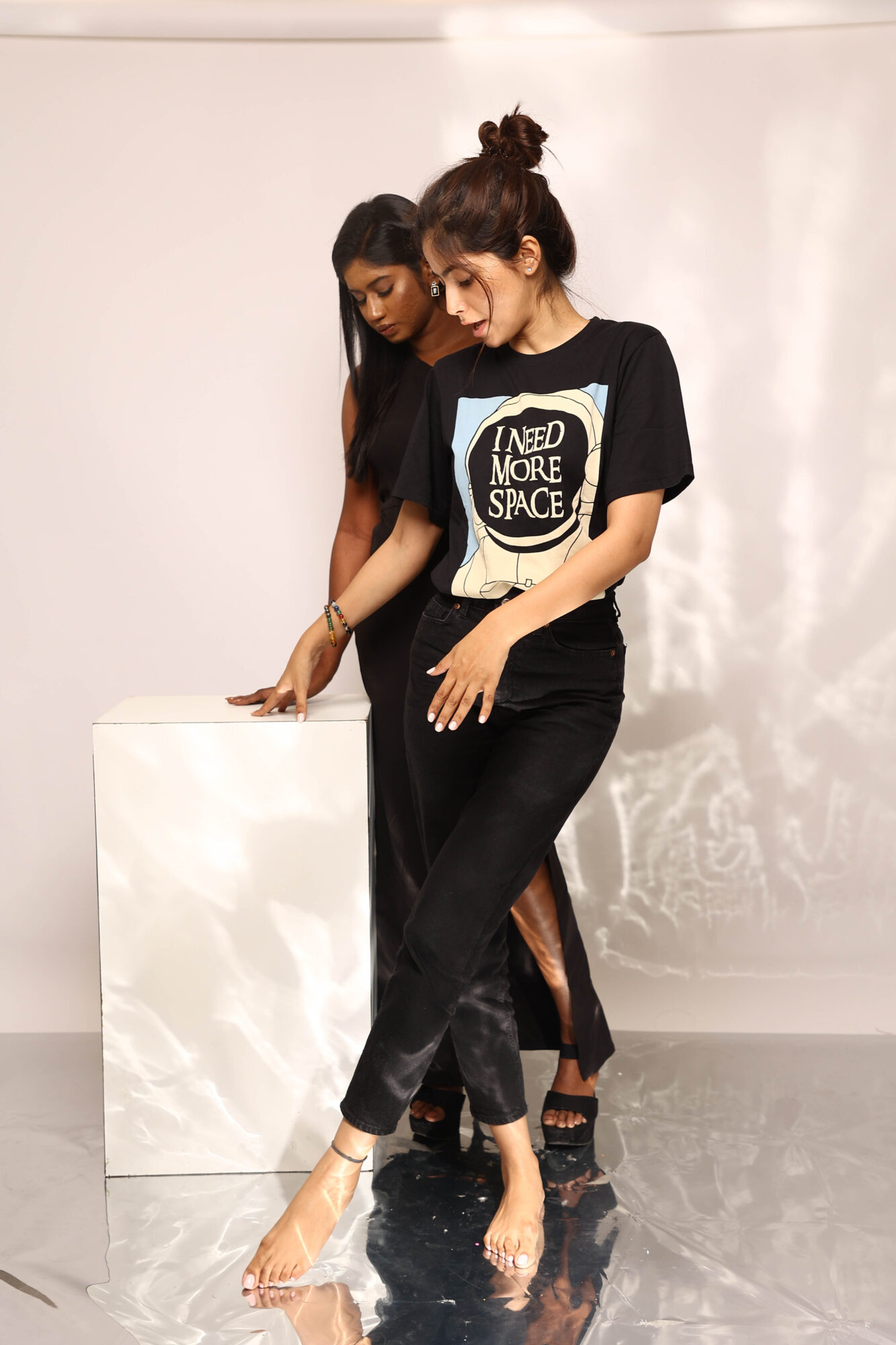 Two women walk barefoot on a reflective surface, one wearing a black t-shirt with a space-themed graphic, the other in black pants.