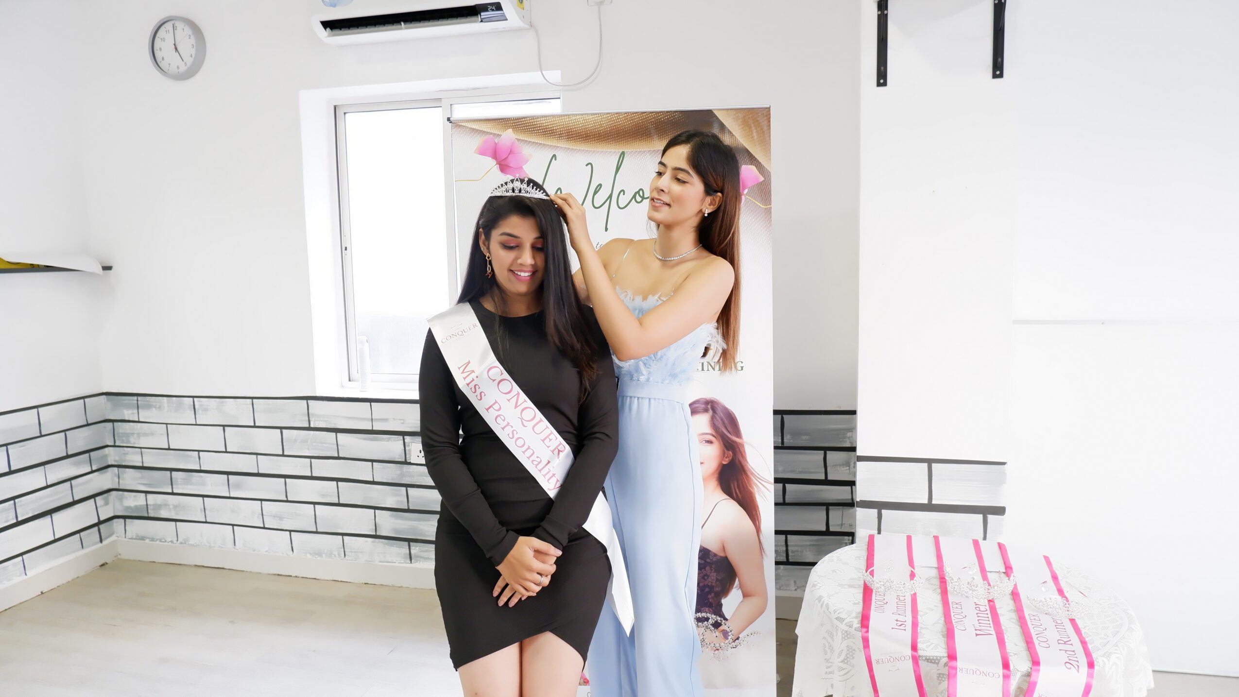 Two women standing indoors, one adjusting the other's hair, with a window and a poster in the background.