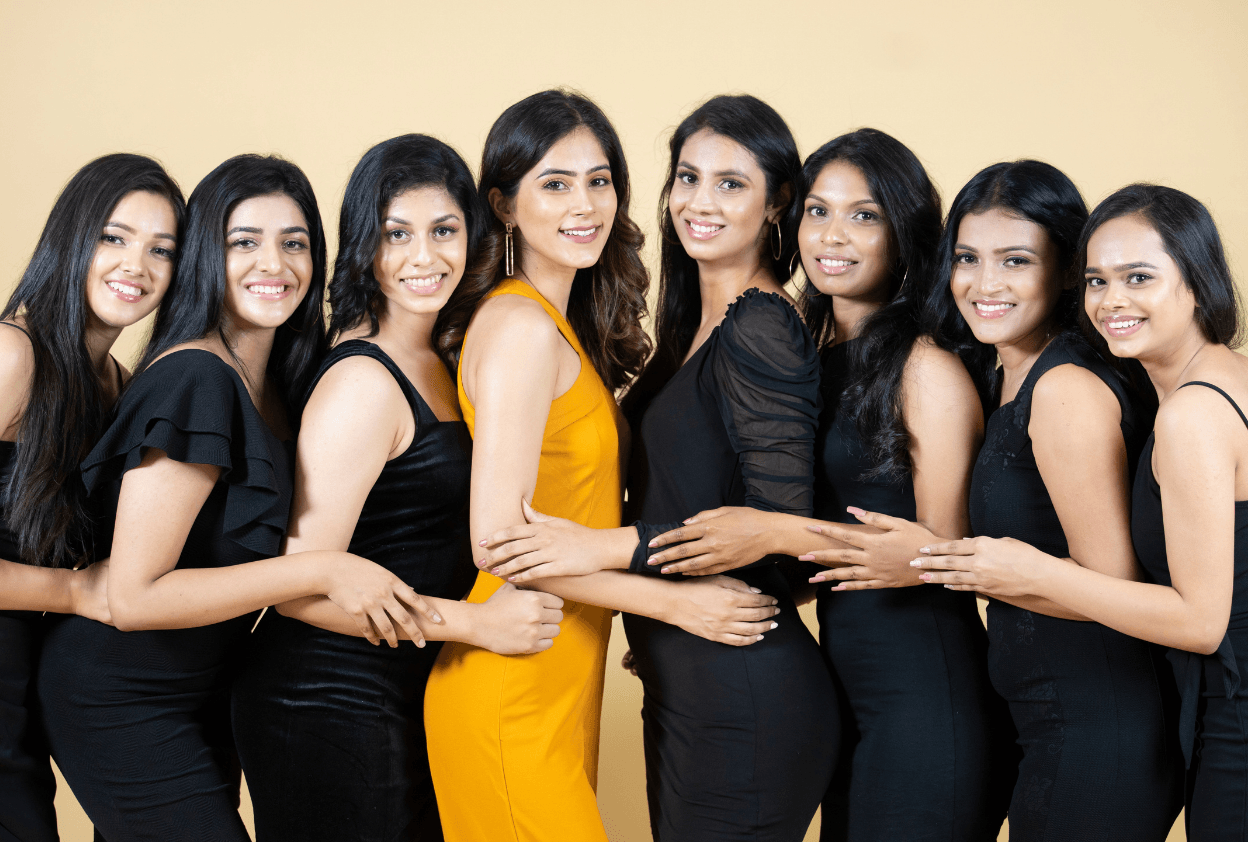 Group of nine women standing together, smiling, with one woman in a yellow dress in the center, against a plain background.